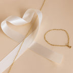 The Heart Link Chain Necklace in 18K Gold, featuring a heart clasp, sits next to a white satin ribbon on a beige background.