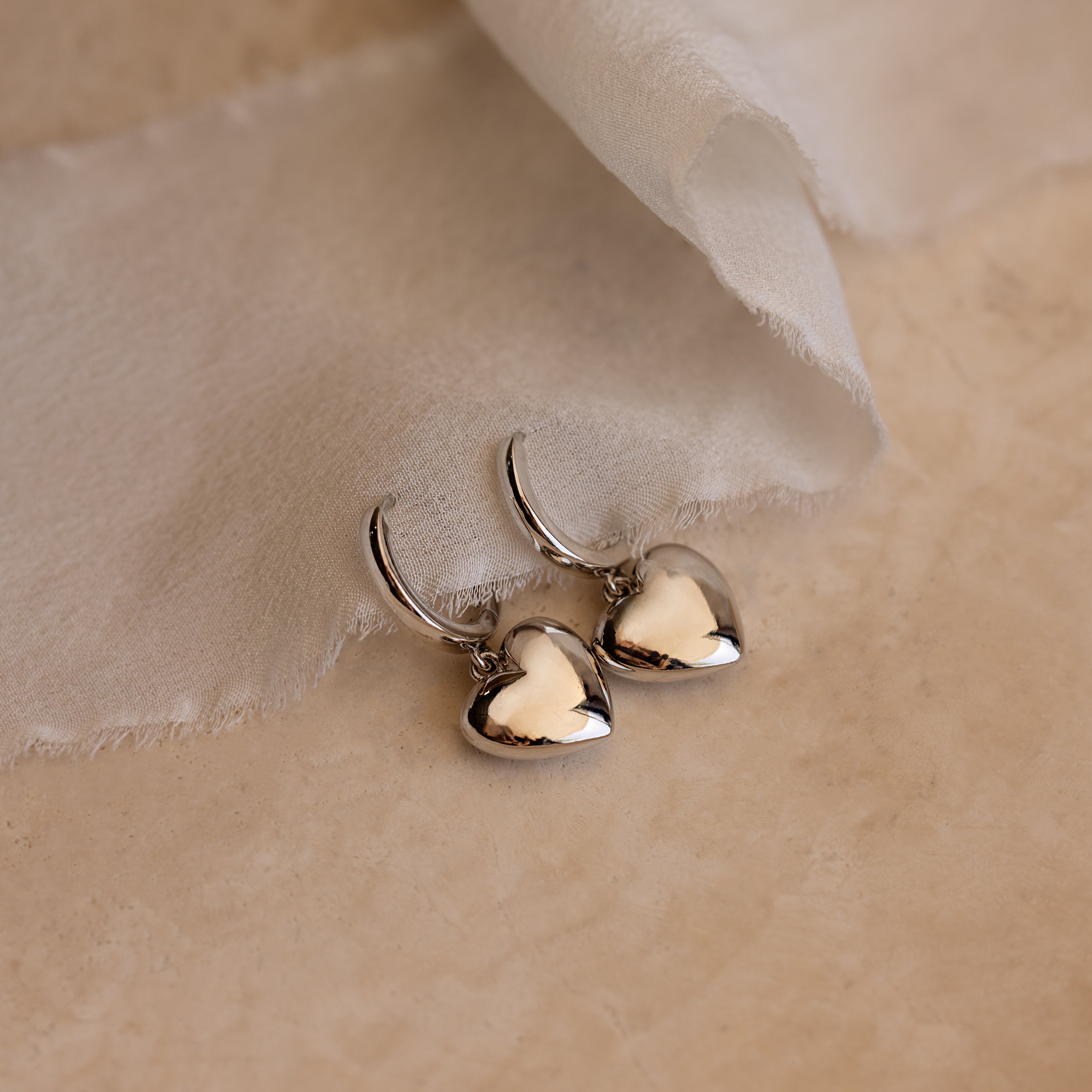 Puff Heart Charm Huggies in Sterling Silver, elegant heart-shaped hoop earrings, are displayed on a light beige surface with soft fabric nearby.