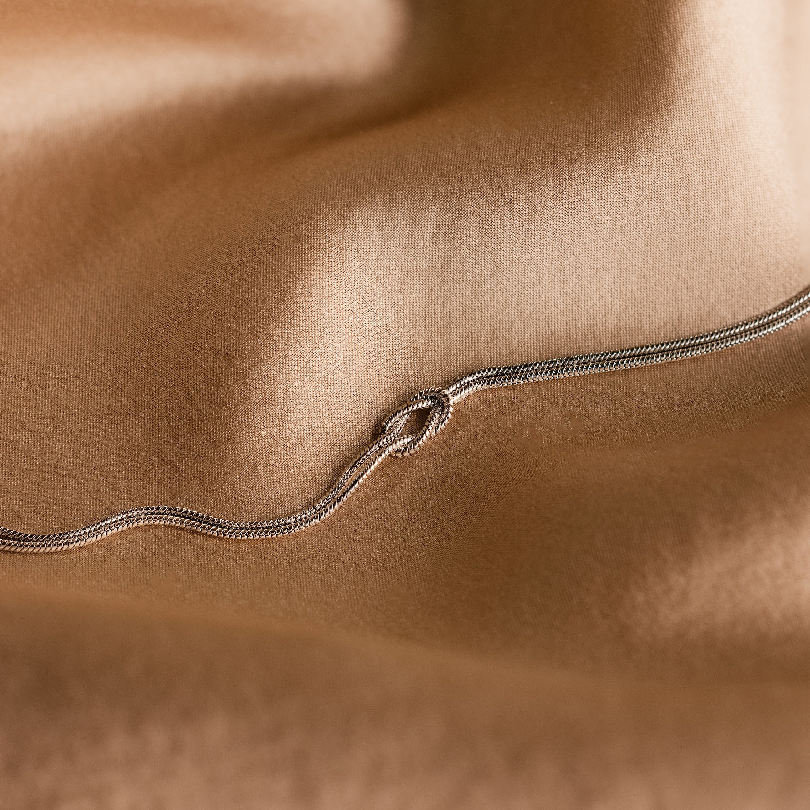 A delicate white gold double chain forms a loose knot on soft, light brown fabric—a perfect Love Knot Bracelet in White Gold gift for couples.