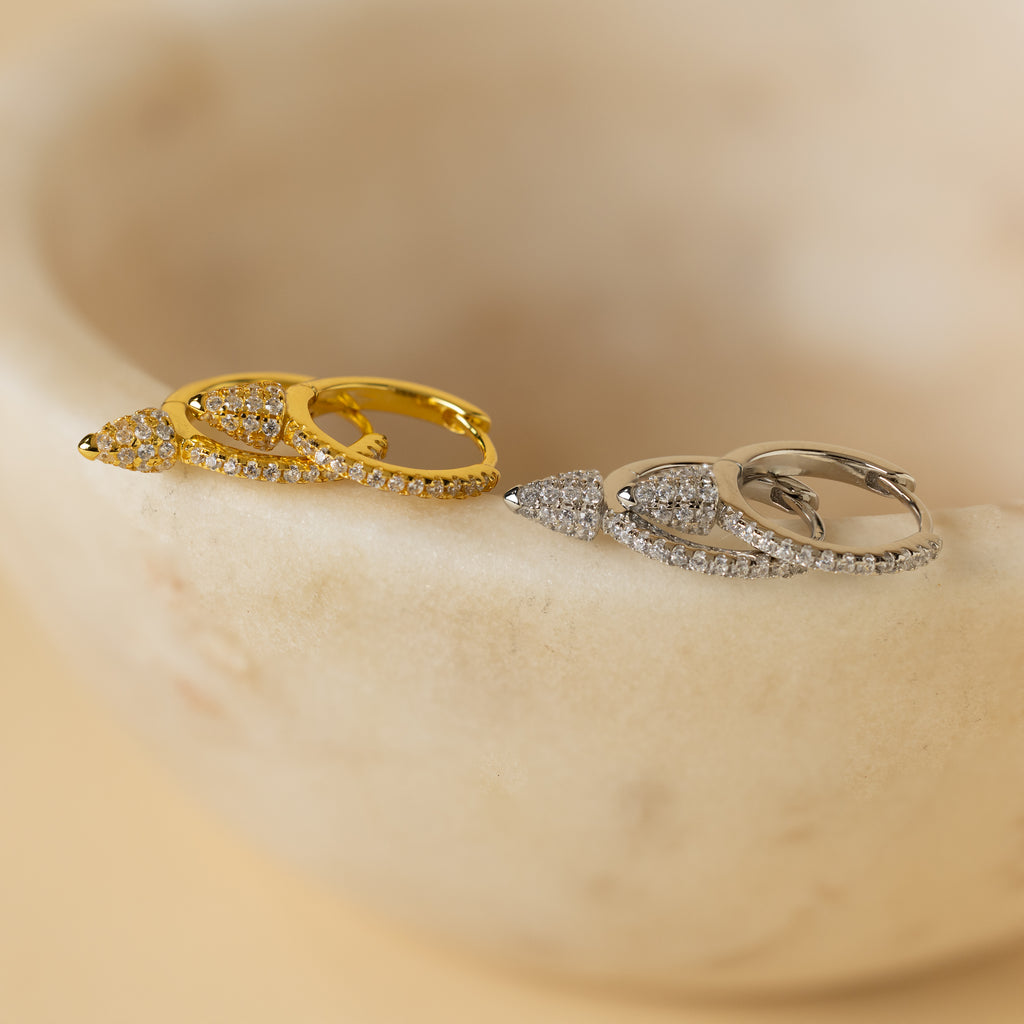 The Pave Spike Huggies, teardrop-shaped earrings in gold and silver with small crystals and an edgy cone design, are elegantly displayed on a smooth beige stone.