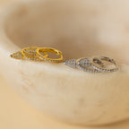 The Pave Spike Huggies, teardrop-shaped earrings in gold and silver with small crystals and an edgy cone design, are elegantly displayed on a smooth beige stone.