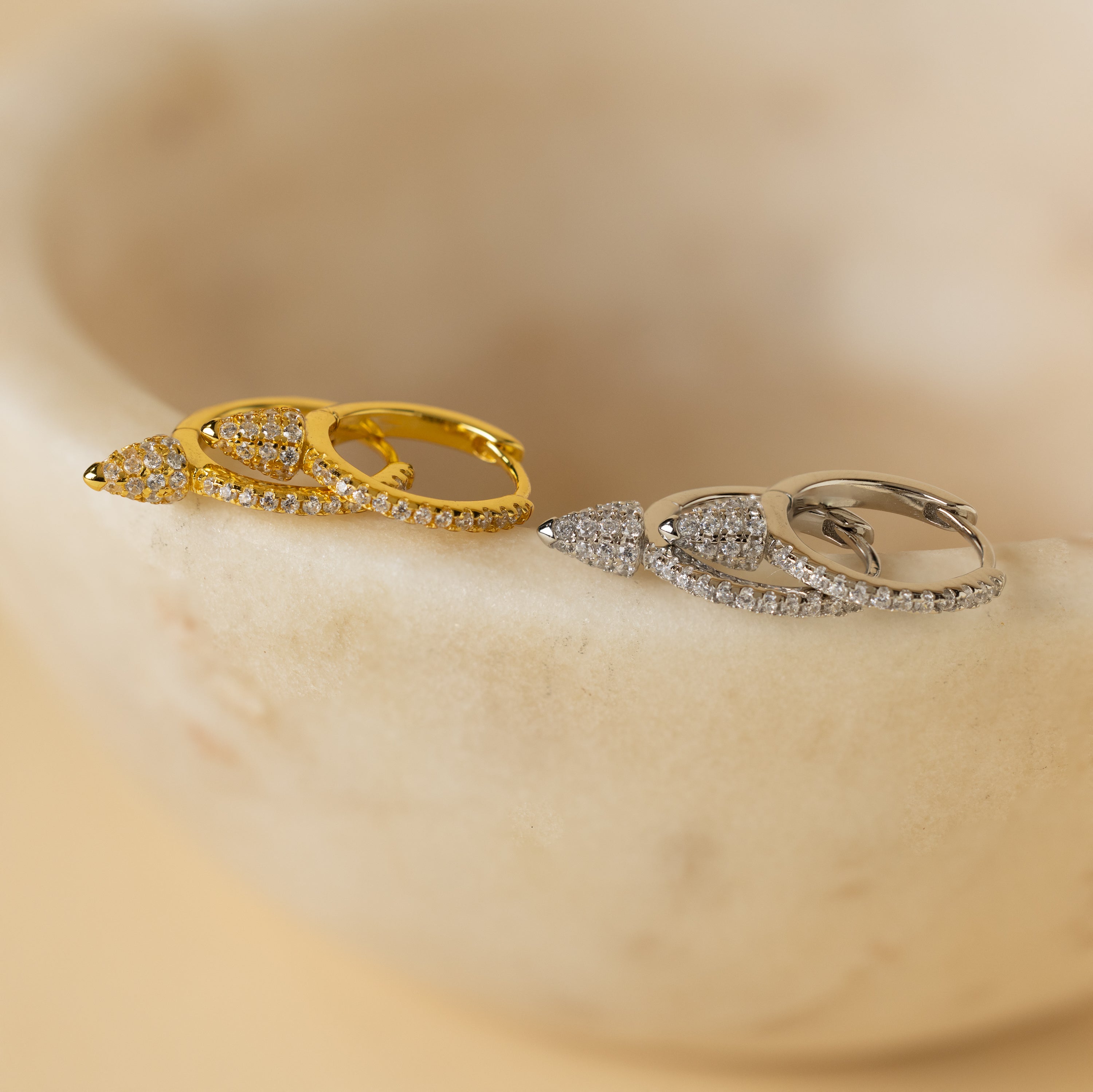 The Pave Spike Huggies, teardrop-shaped earrings in gold and silver with small crystals and an edgy cone design, are elegantly displayed on a smooth beige stone.