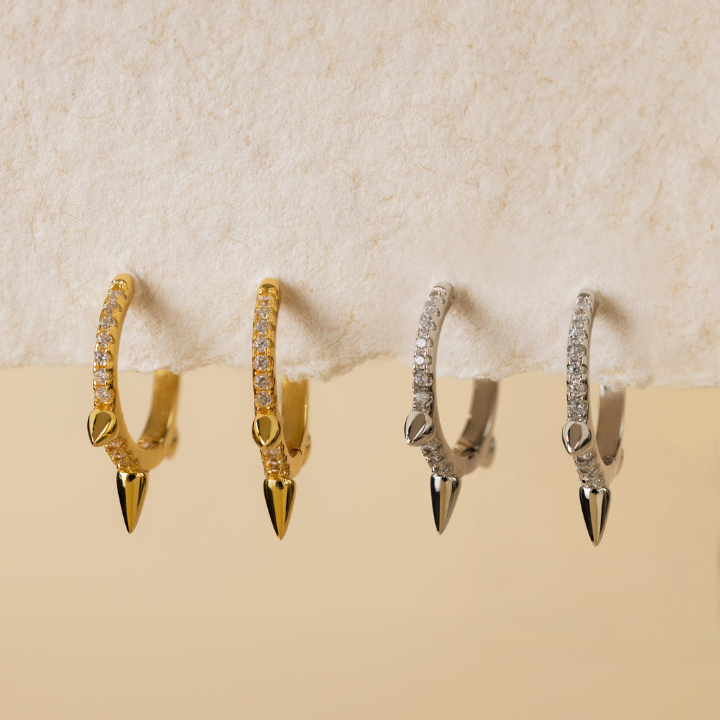 Two pairs of Pave Triple Spike Huggies—diamond hoop earrings with pavé stones and spike charms, in gold and silver—rest on a textured surface, ideal for minimalist jewelry lovers.