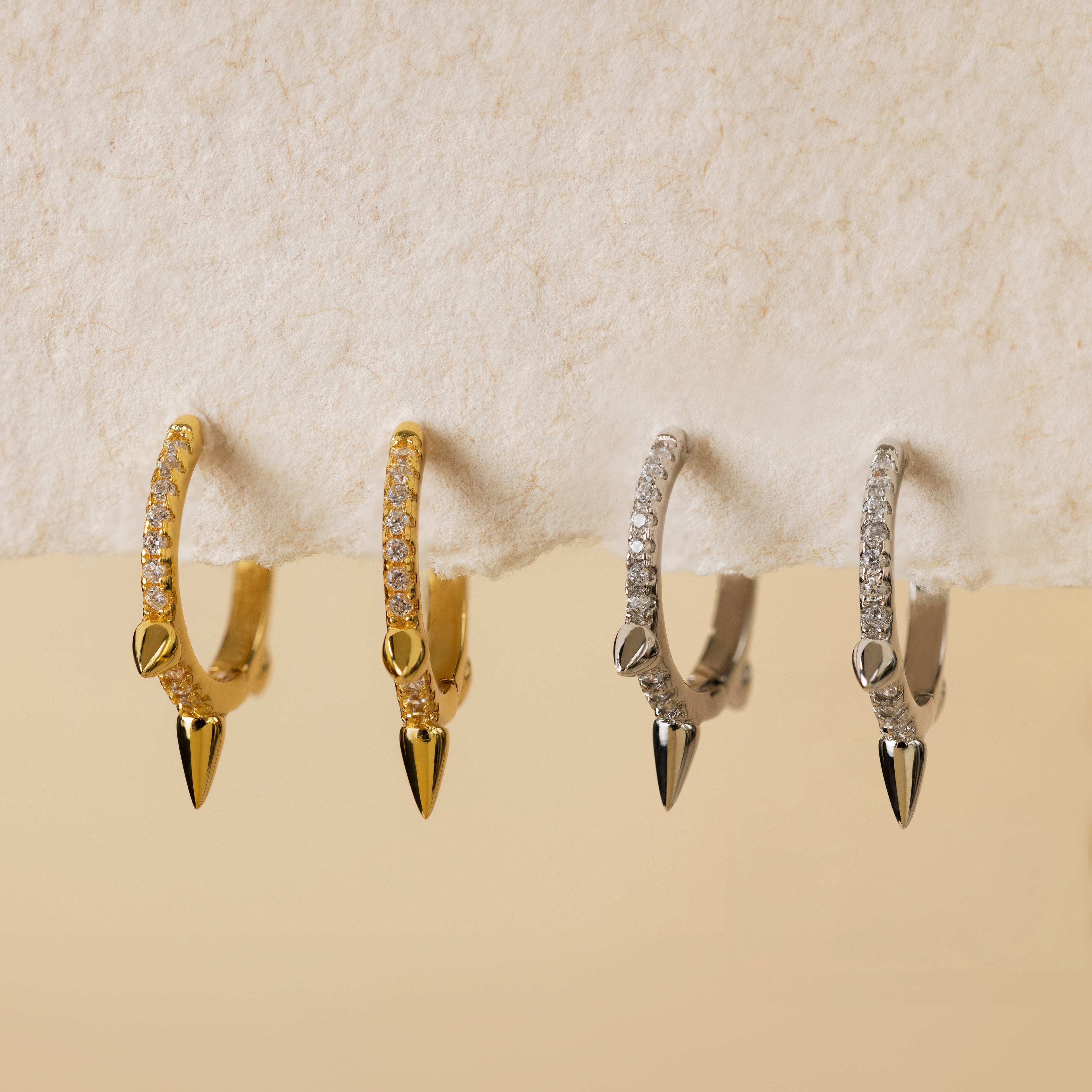 Two pairs of Pave Triple Spike Huggies—diamond hoop earrings with pavé stones and spike charms, in gold and silver—rest on a textured surface, ideal for minimalist jewelry lovers.