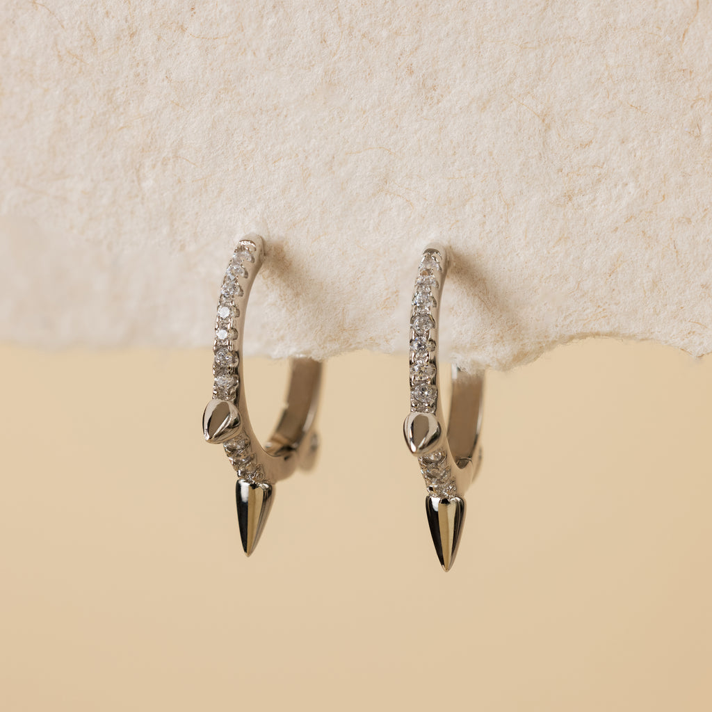 Minimalist jewelry: Pave Triple Spike Huggies—silver hoop earrings with small clear stones and pointed tips, shown on textured off-white paper.