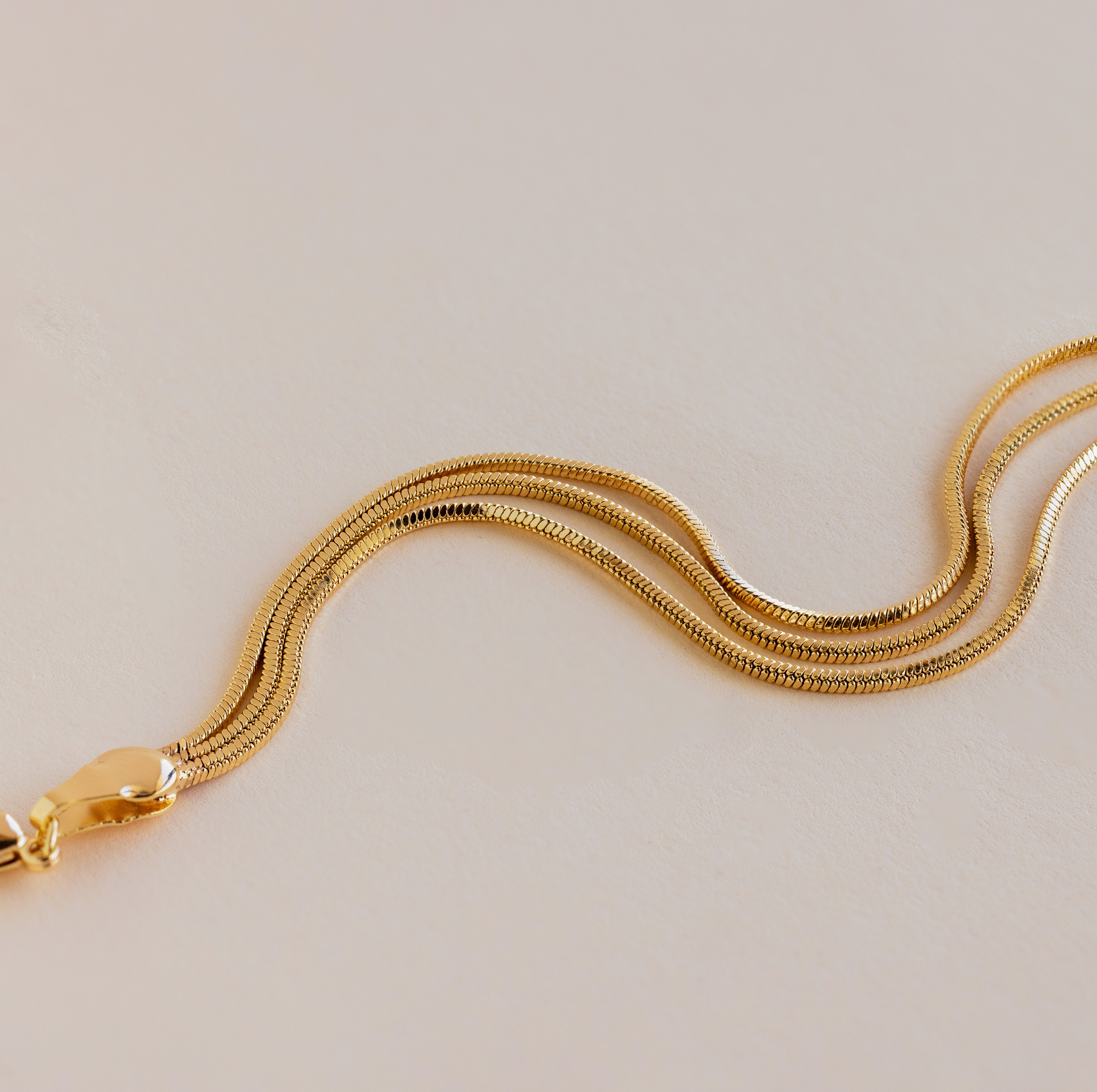 A close-up of the Triple Snake Chain Bracelet in 18K Gold with a lobster clasp on a light beige background, perfect for layering or pairing with minimal jewelry.