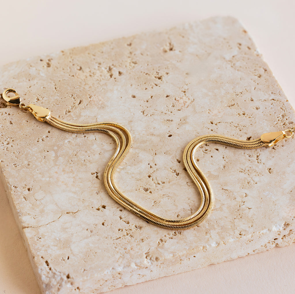 The Triple Snake Chain Bracelet in 18K Gold features lobster clasps and is perfect for a minimal jewelry collection, showcased on a textured stone surface for a chic layered style.