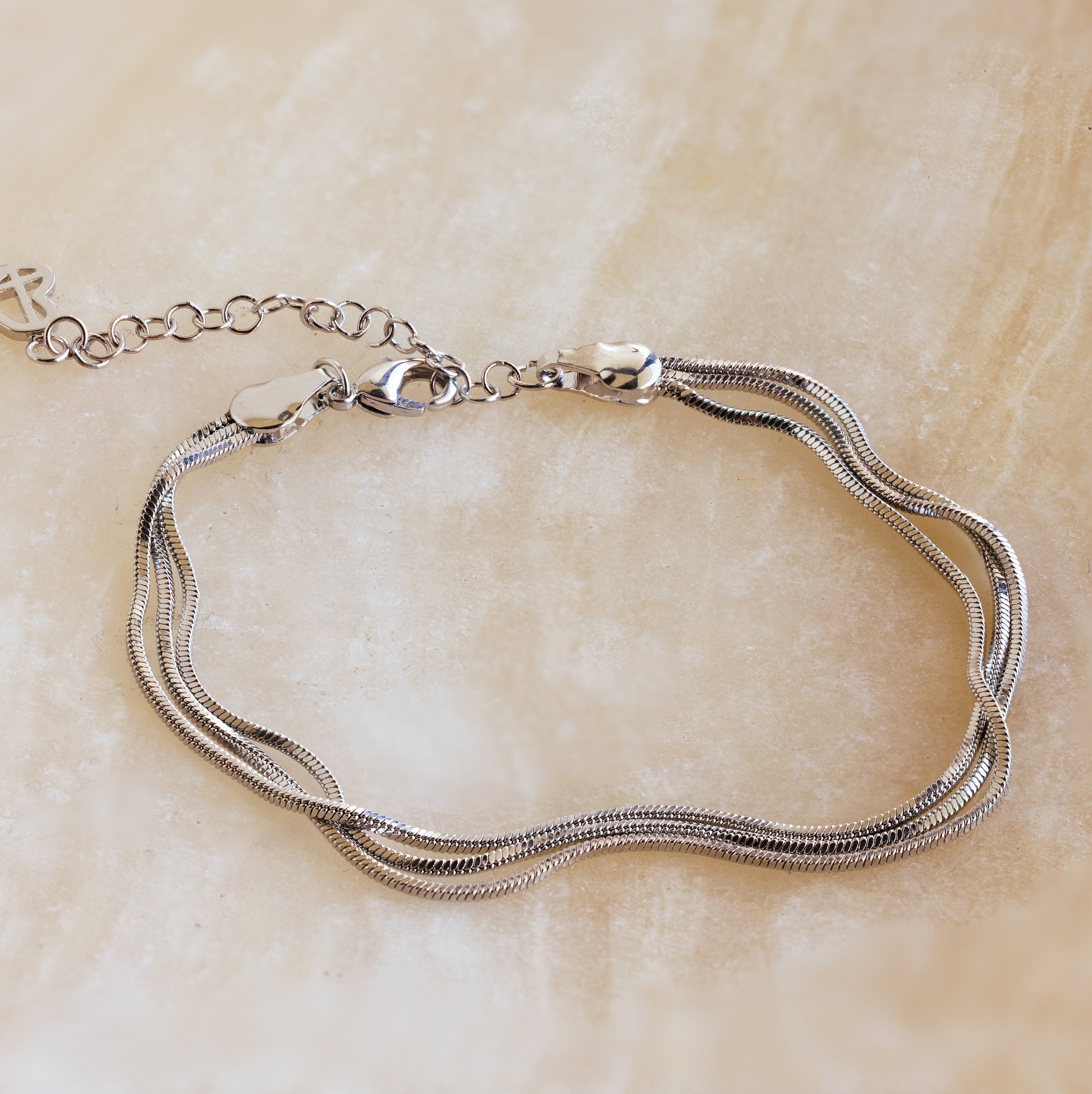 The Triple Snake Chain Bracelet in White Gold, featuring a lobster clasp, is displayed on a light beige surface—ideal for adding a layered touch to your minimal jewelry collection.