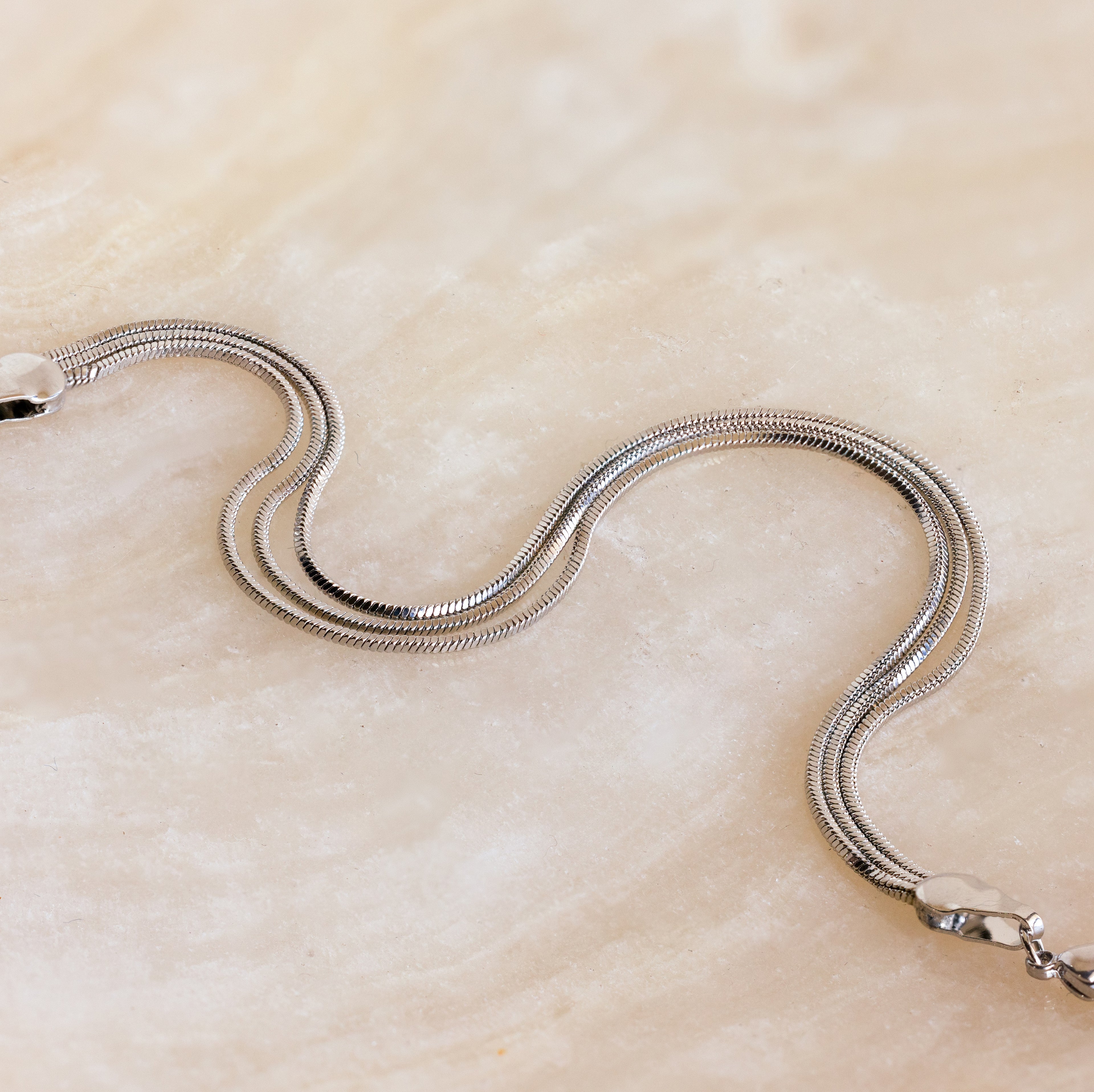 A minimal jewelry piece, the Triple Snake Chain Bracelet in White Gold lies in a gentle curve on a marble-like surface, perfect for chic layered looks.