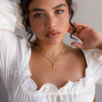 Woman with curly hair wears a white ruffled blouse and layered gold necklaces, including the Pearl Initial Tag Necklace—a customizable chain that's the perfect Christmas gift—posing with her hands near her face.