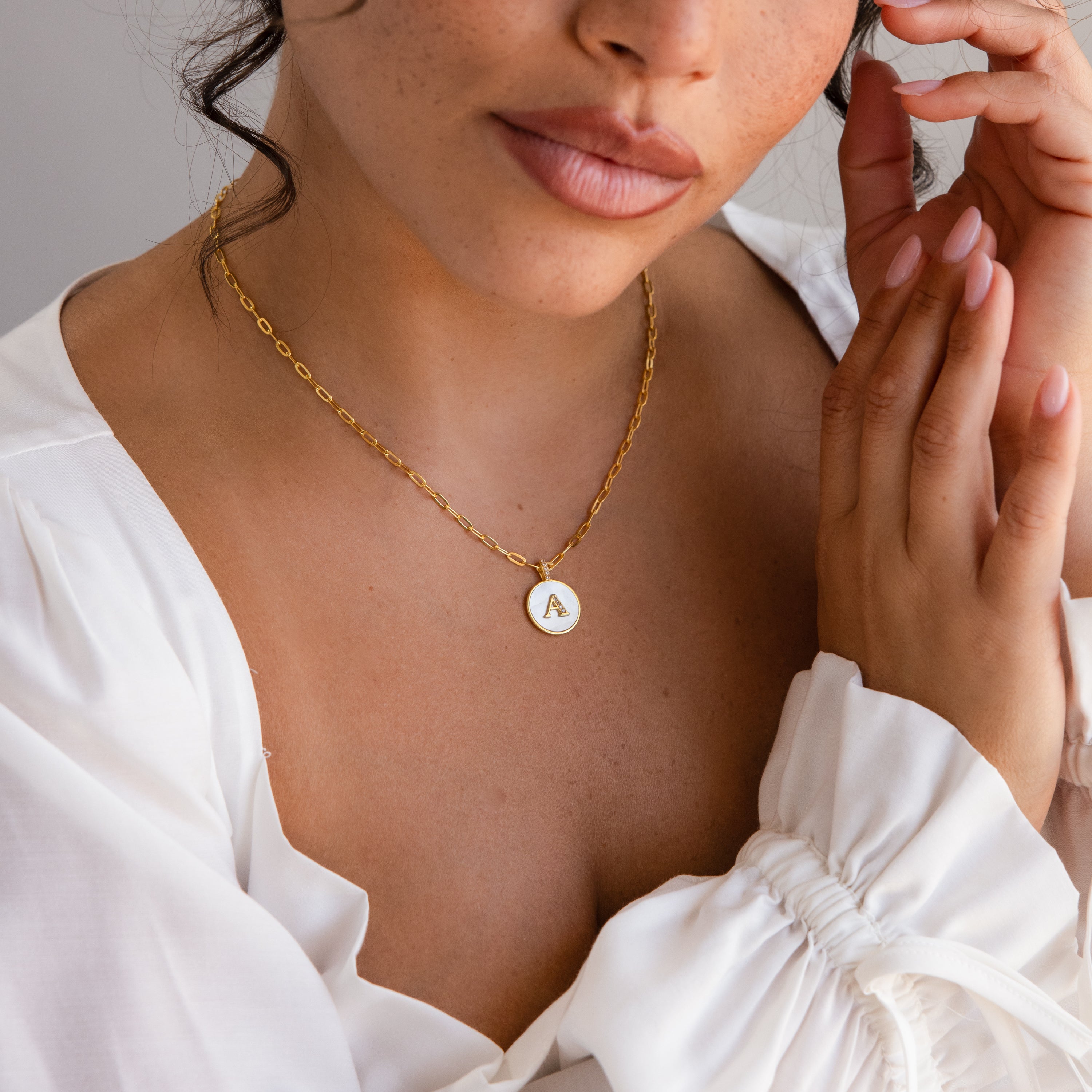 Woman wearing the Pearl Initial Disc Necklace with a gold A pendant and a white blouse, hands gently touching her face.
