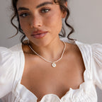 A woman with wavy hair wears a white top and the Pearl Initial Disc Necklace, which features a round pendant engraved with the letter K.