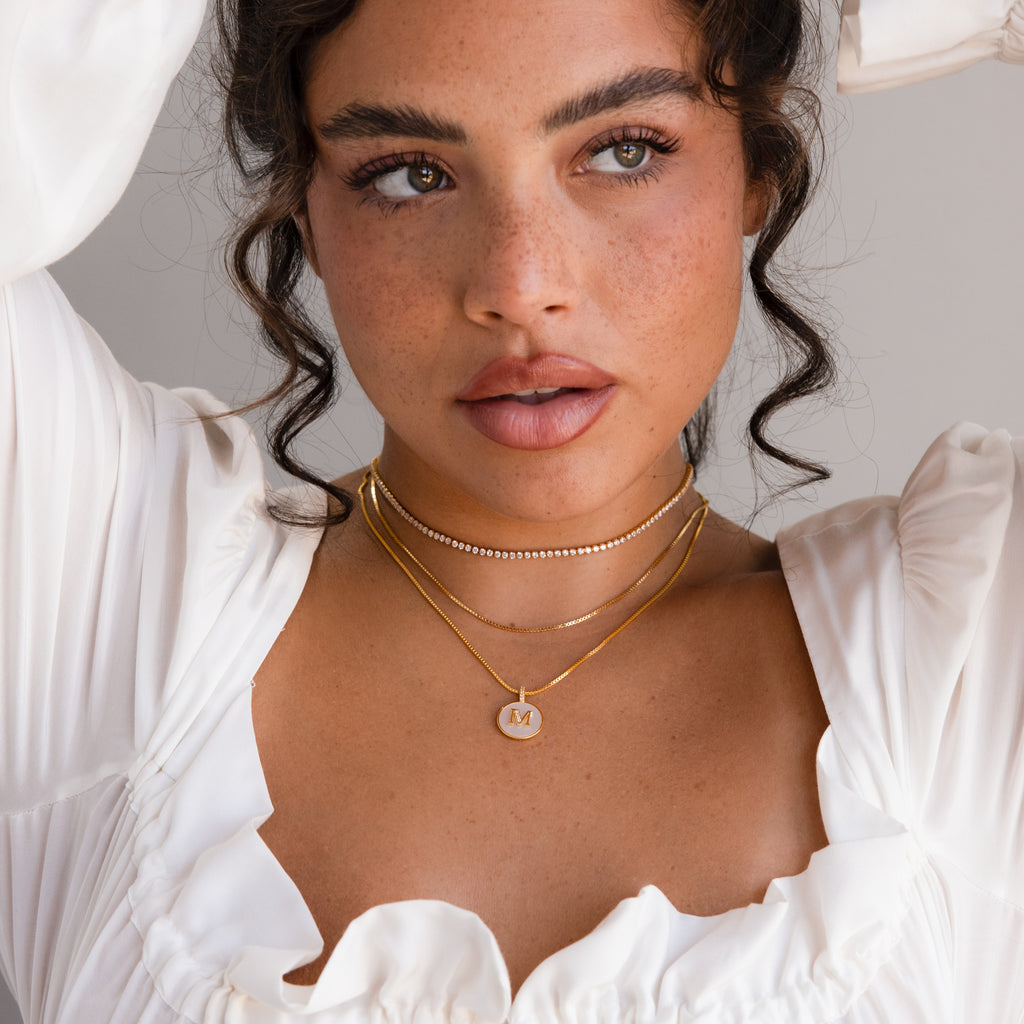 Woman with curly hair in a white top, wearing layered gold necklaces including the Pearl Initial Disc Necklace, looking slightly to the side.