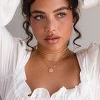 Woman with curly hair in a white top, wearing layered gold necklaces including the Pearl Initial Disc Necklace, looking slightly to the side.