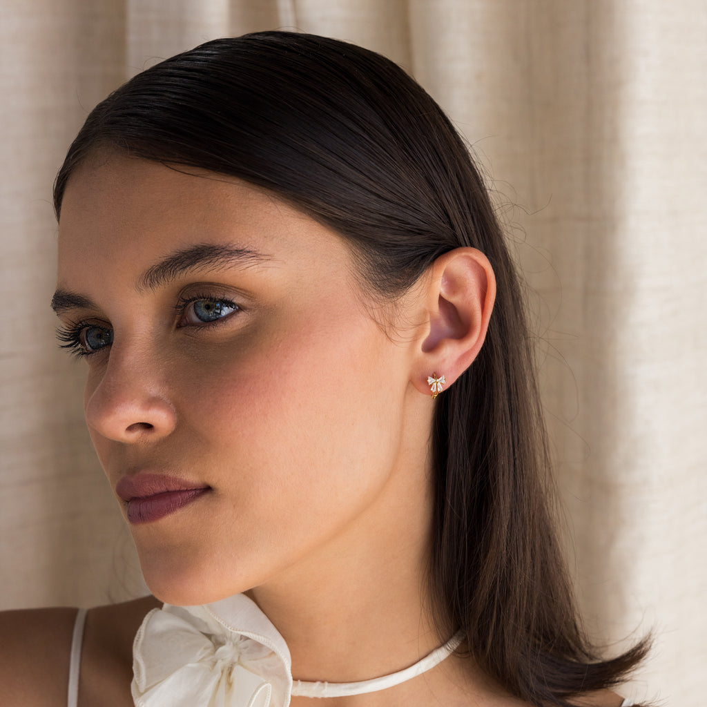 Woman with straight dark hair wears a white bow top and Diamond Ribbon Bow Huggies in 18K Gold, standing by beige curtains.
