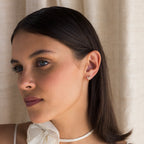 Woman with straight dark hair wears a white bow top and Diamond Ribbon Bow Huggies in 18K Gold, standing by beige curtains.