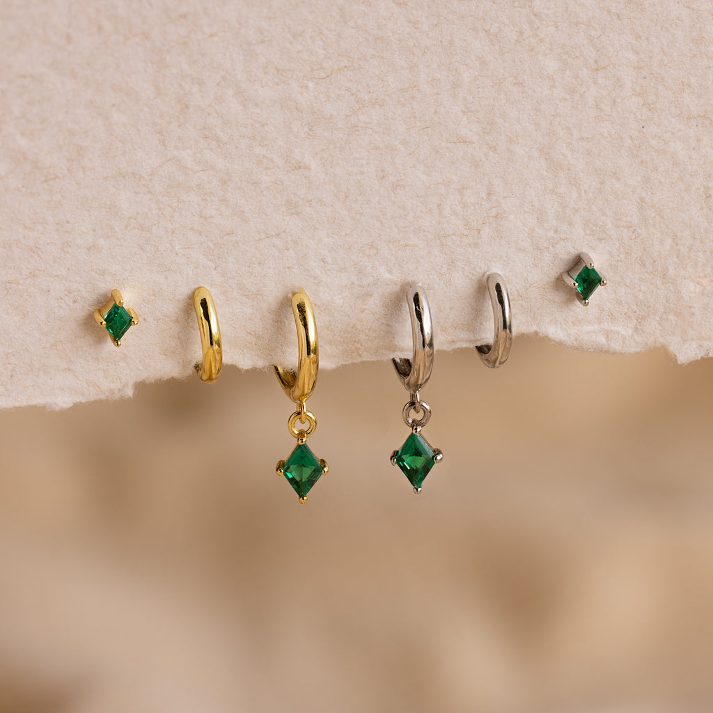 The Emerald Earrings Set features five green emerald earrings, two gold hoops, two silver hoops, and one small stud displayed on textured paper.