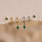 The Emerald Earrings Set features five green emerald earrings, two gold hoops, two silver hoops, and one small stud displayed on textured paper.
