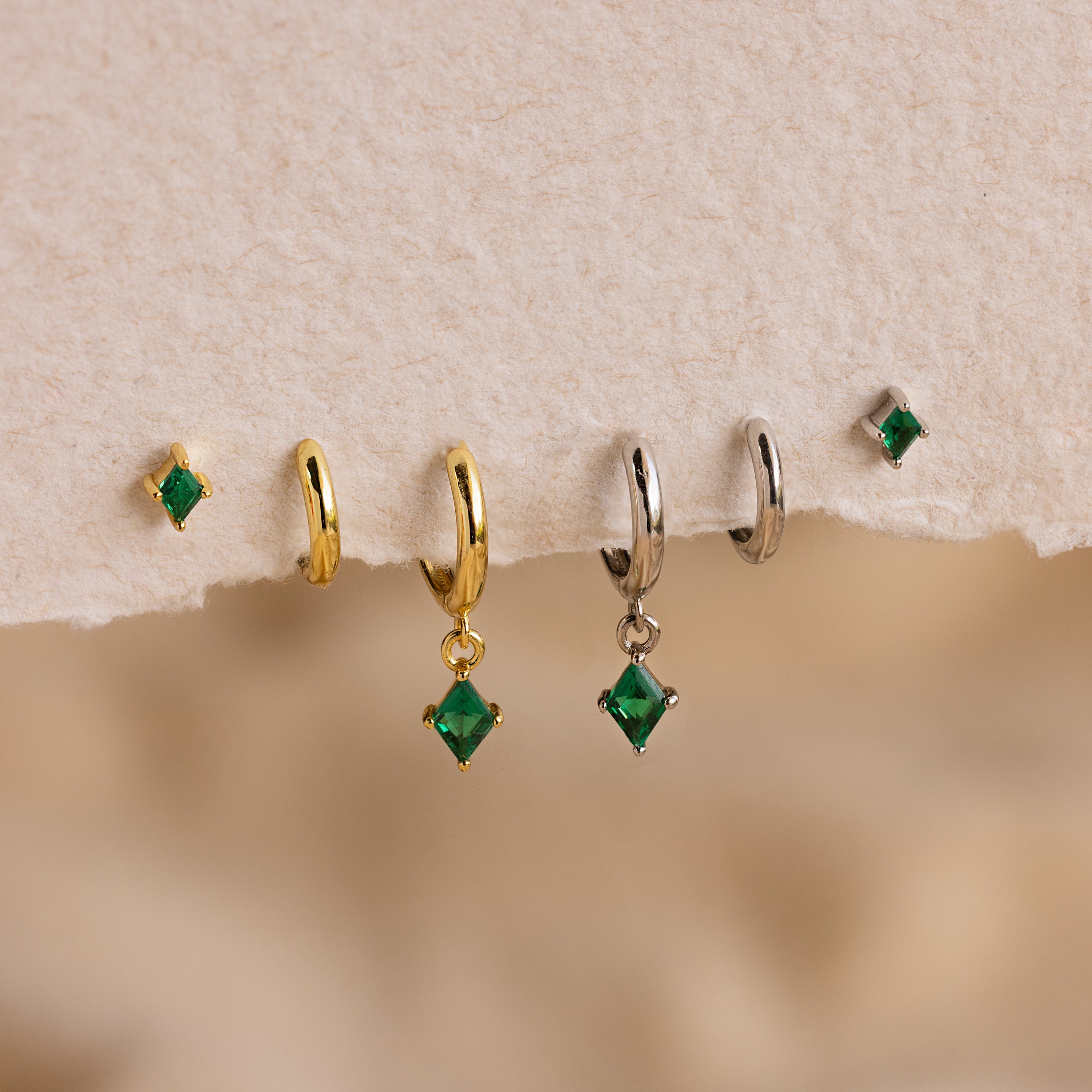 The Emerald Earrings Set features five green emerald earrings, two gold hoops, two silver hoops, and one small stud displayed on textured paper.