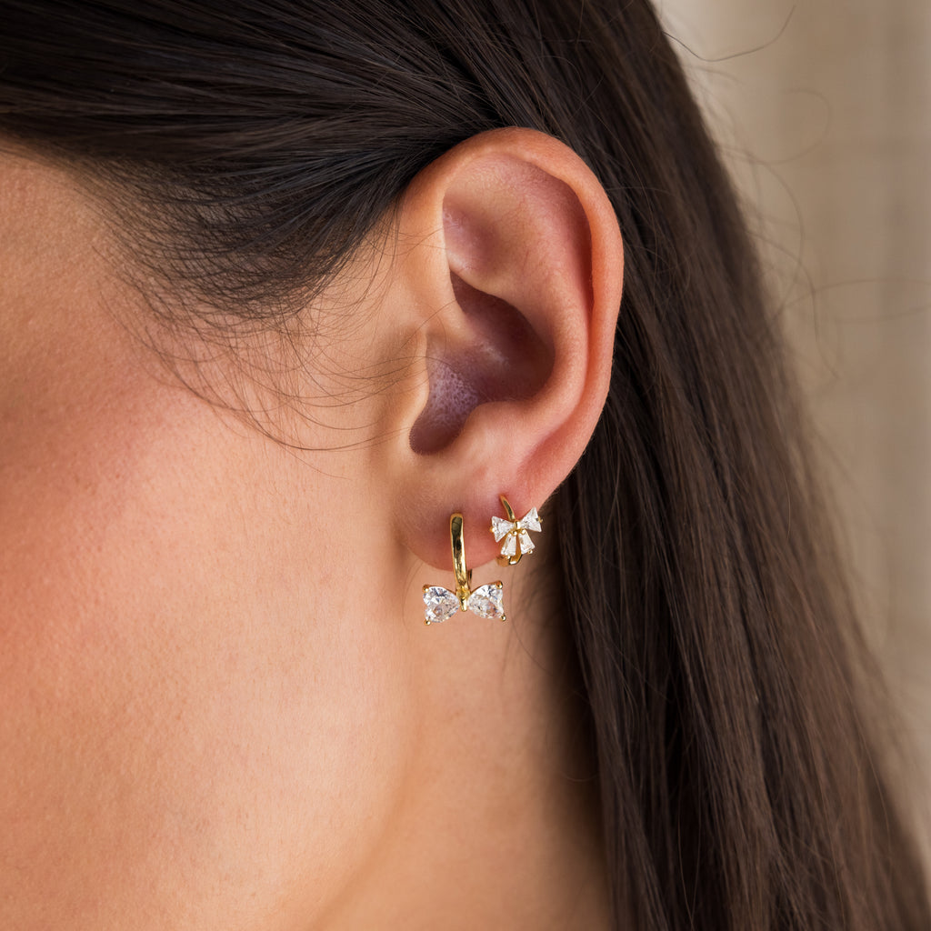 Close-up of a womans ear wearing two gold earrings: Diamond Bow Huggies and a dainty flower stud.