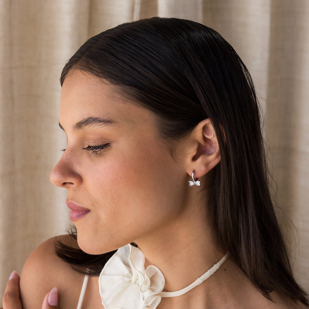 Woman with straight dark hair wears Diamond Bow Huggies, butterfly earrings, and a white flower choker, looking to the left.