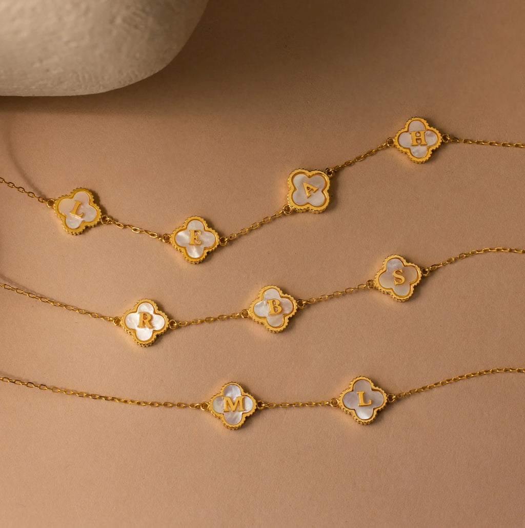 Three Gemstone Clover Letter Necklaces with white inlays and engraved initials—L, H, R, B, S, M, and L—displayed on a beige surface. Each pairs beautifully with personalized initial necklaces or other 925 silver pieces.