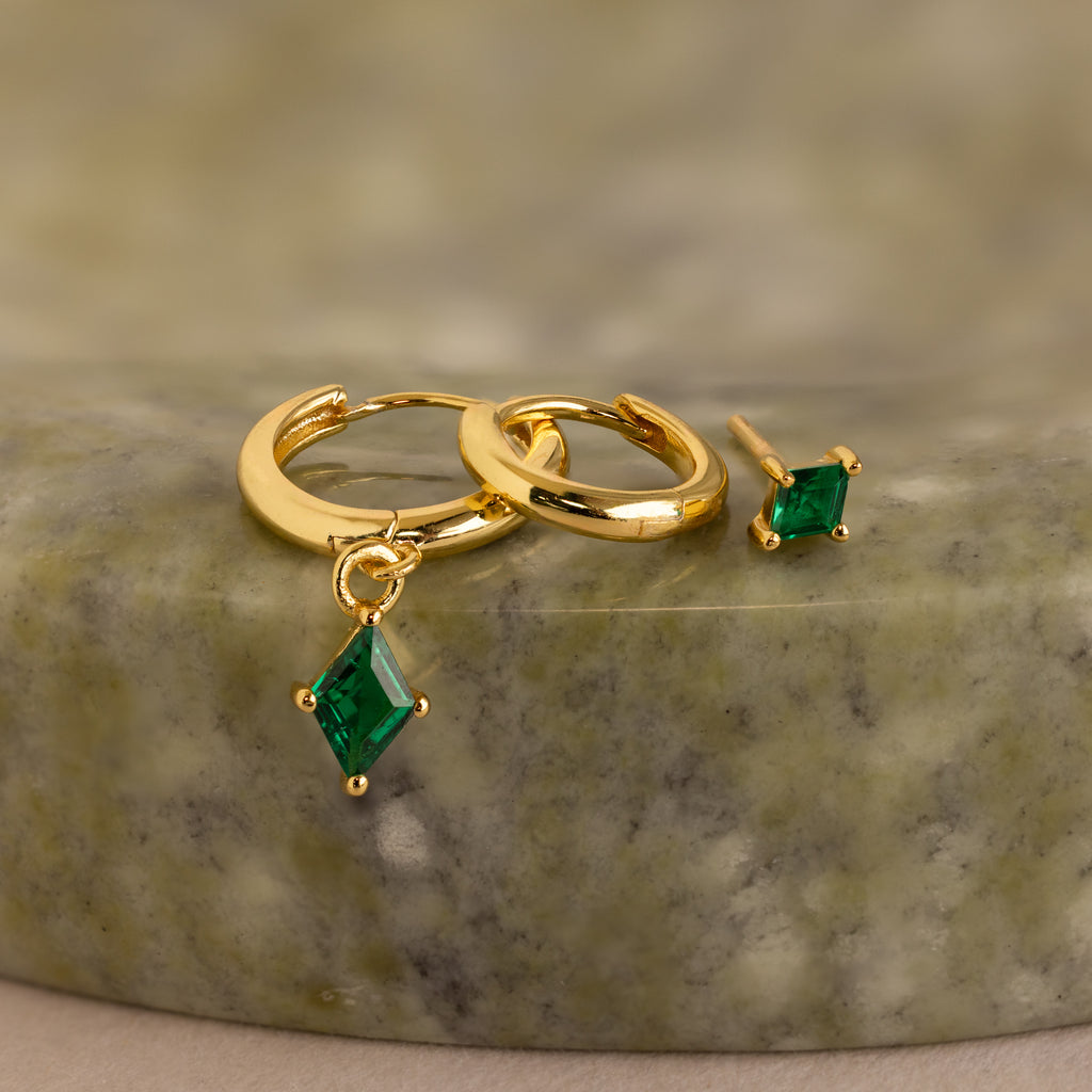 The Emerald Earrings Set features gold hoops with green emerald charms and a matching gemstone stud, elegantly displayed on a green stone surface.