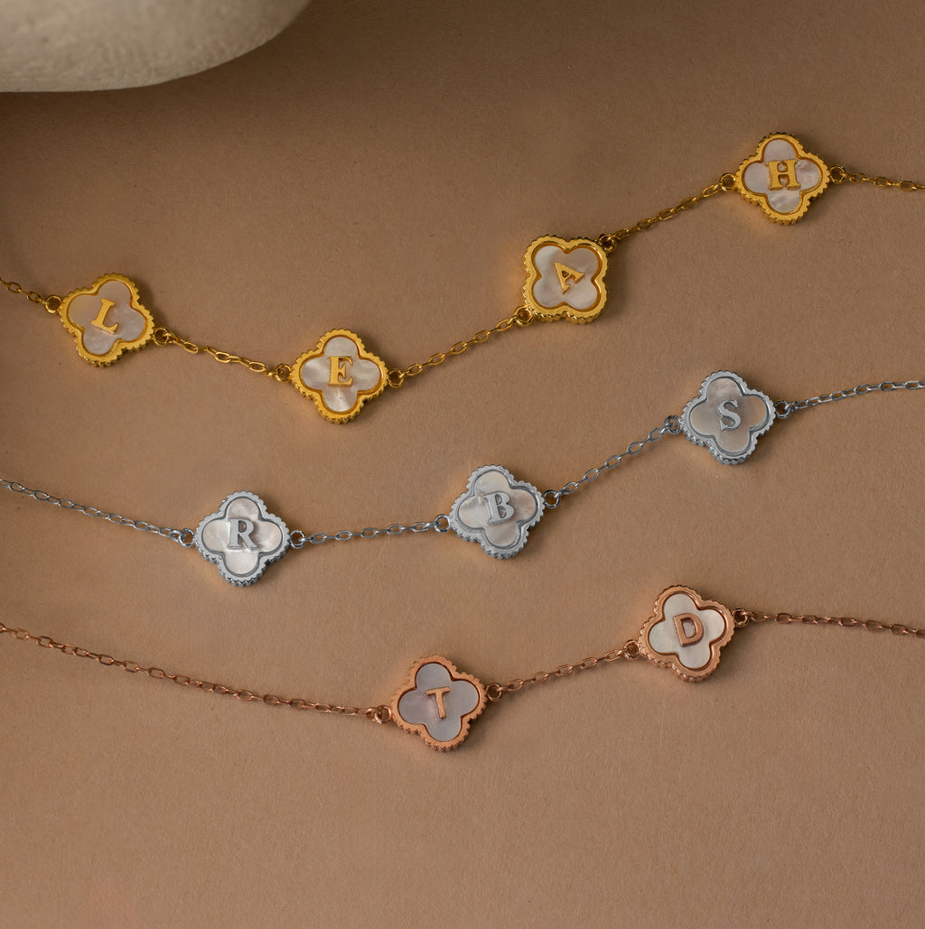 Three Pearl Clover Letter Necklaces, each with clover-shaped charms and the letters L E A H, R B S, and T D, are displayed on a brown background, showcasing their personalized elegance.