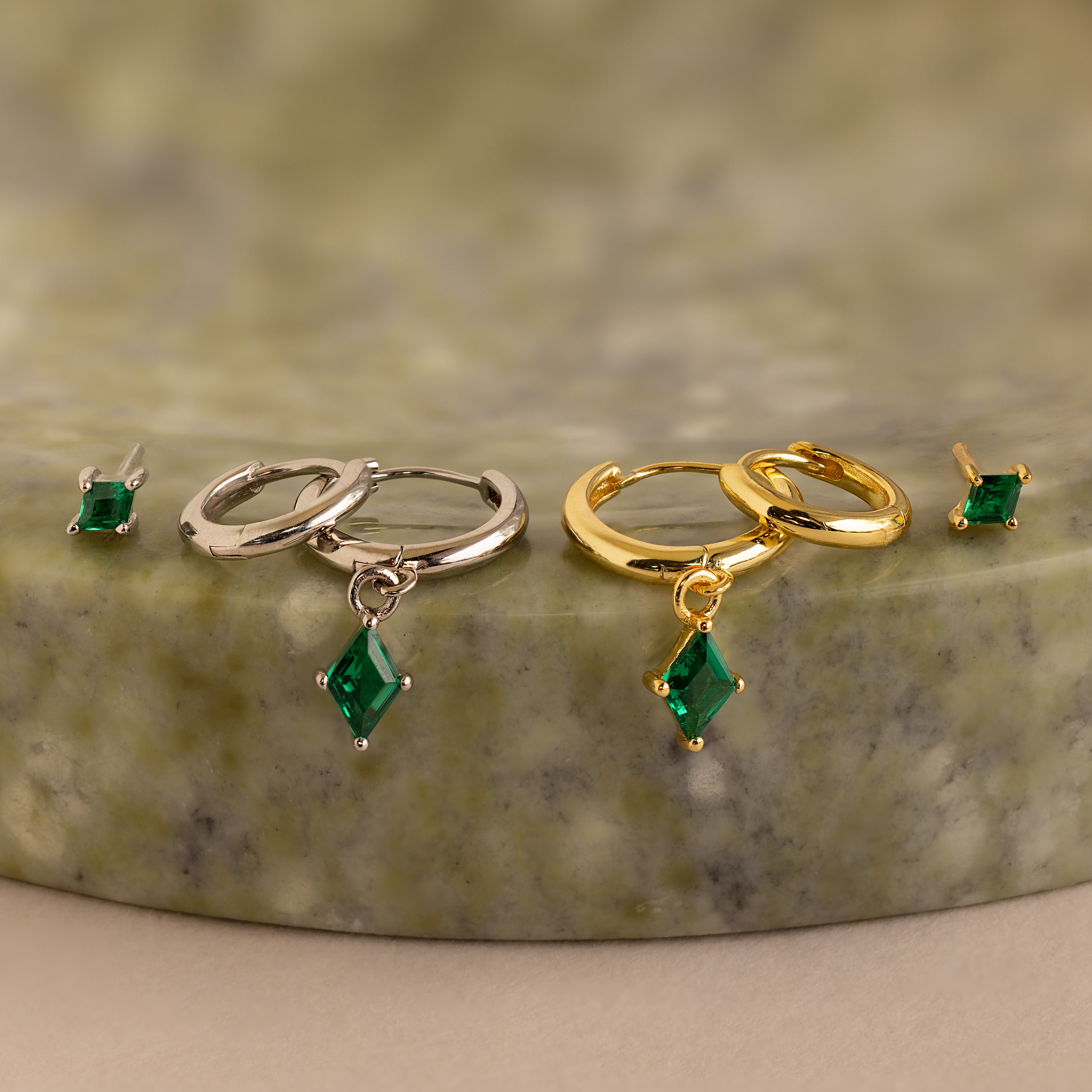 The Emerald Earrings Set features two pairs of hoop earrings—one silver and one gold—with green emerald gemstone drops, elegantly displayed on a marble surface.