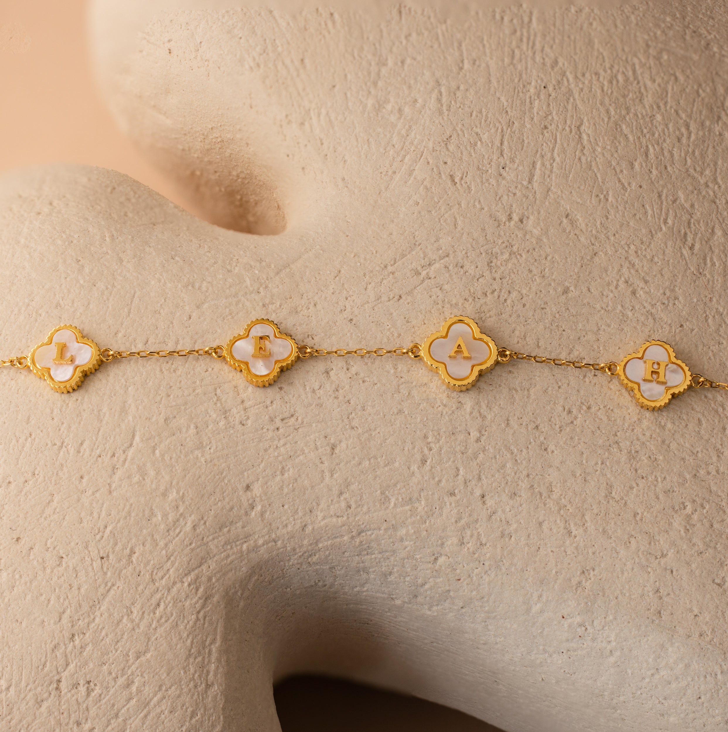 The Pearl Clover Letter Necklace features four clover-shaped charms, each with a white letter—L, F, A, and H—on a textured gold surface, blending personalization with the iconic four-leaf clover design.