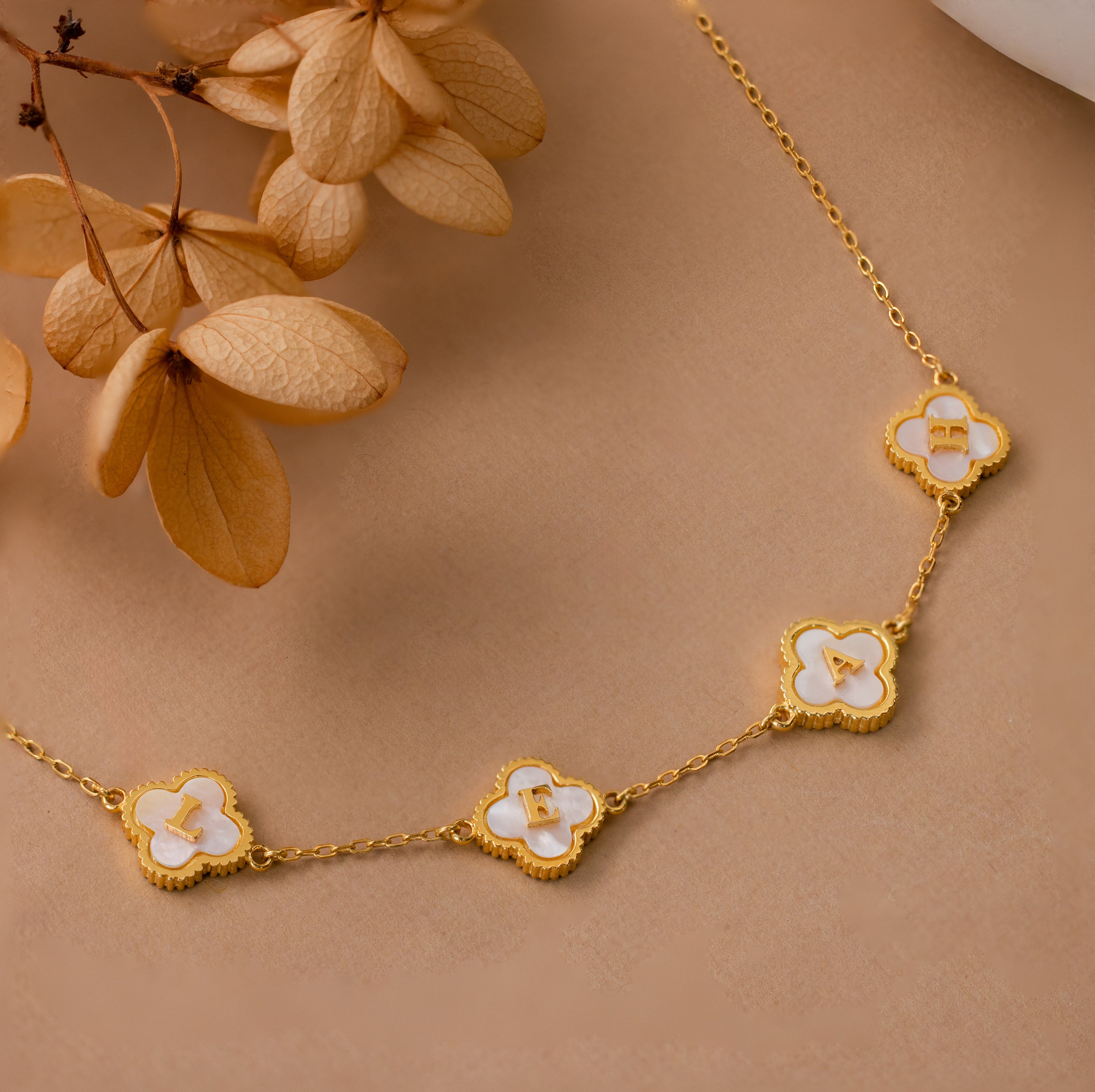 The Pearl Clover Letter Necklace features five white clover-shaped charms, beautifully displayed on a beige surface with dried flowers.