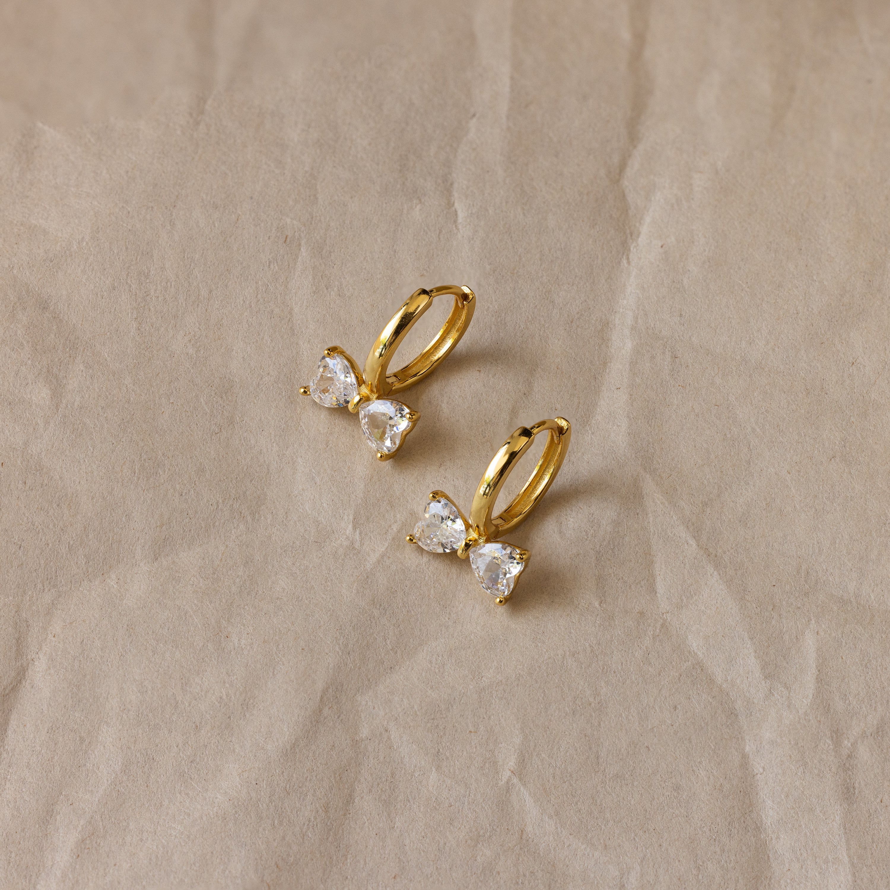 Diamond Bow Huggies: gold hoop earrings adorned with butterfly-shaped crystal charms, displayed elegantly on crumpled beige paper.