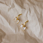 Diamond Bow Huggies in 18K Gold, adorned with two heart-shaped clear stones, rest on crumpled beige paper—an elegant update to classic diamond earrings.