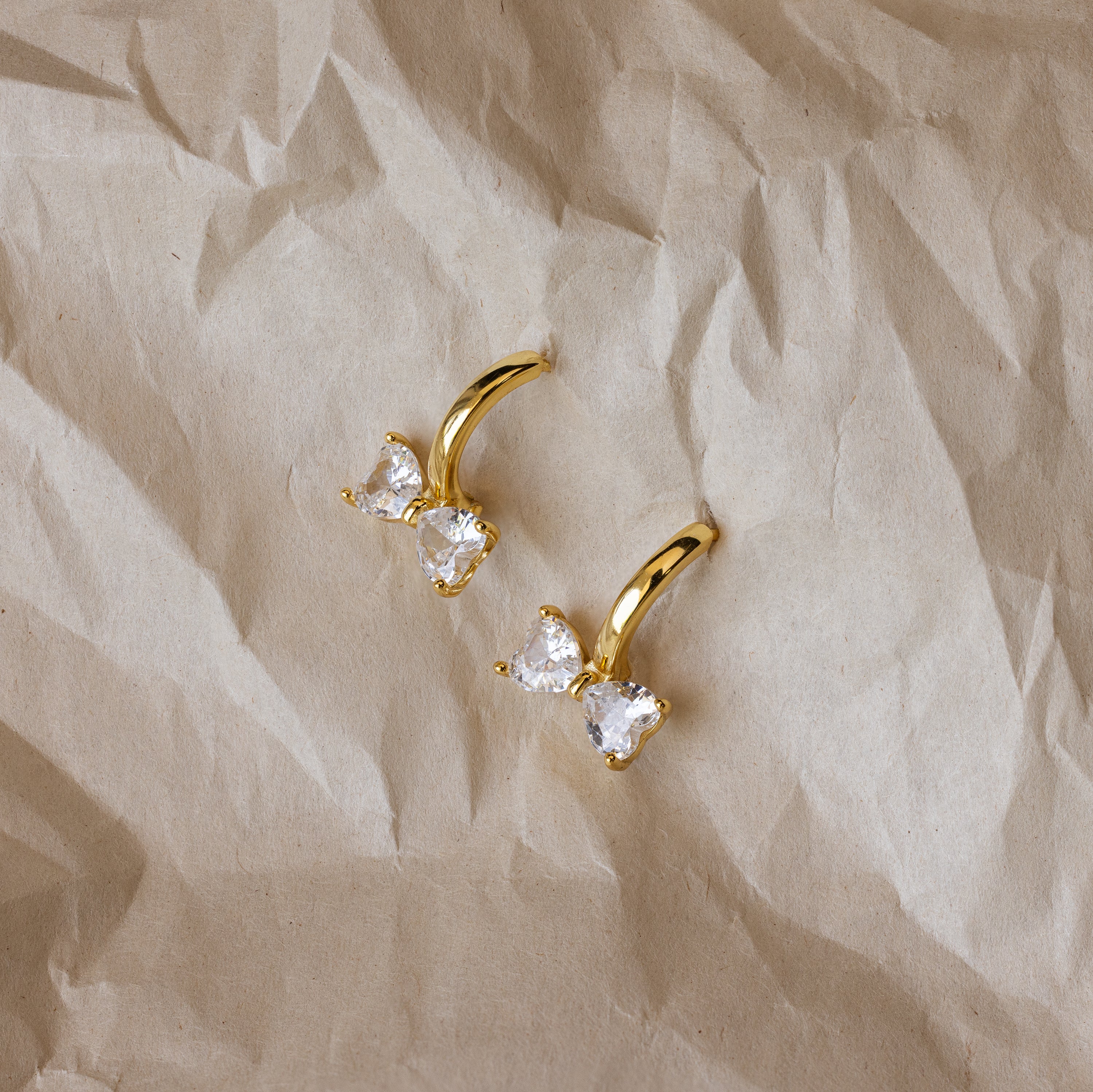 Diamond Bow Huggies in 18K Gold, adorned with two heart-shaped clear stones, rest on crumpled beige paper—an elegant update to classic diamond earrings.