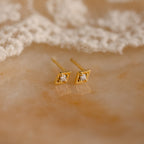 Ericka Diamond Studs are vintage-inspired, gold geometric earrings with small gemstones. These minimalist diamond studs are displayed elegantly on a marble surface next to delicate lace fabric.