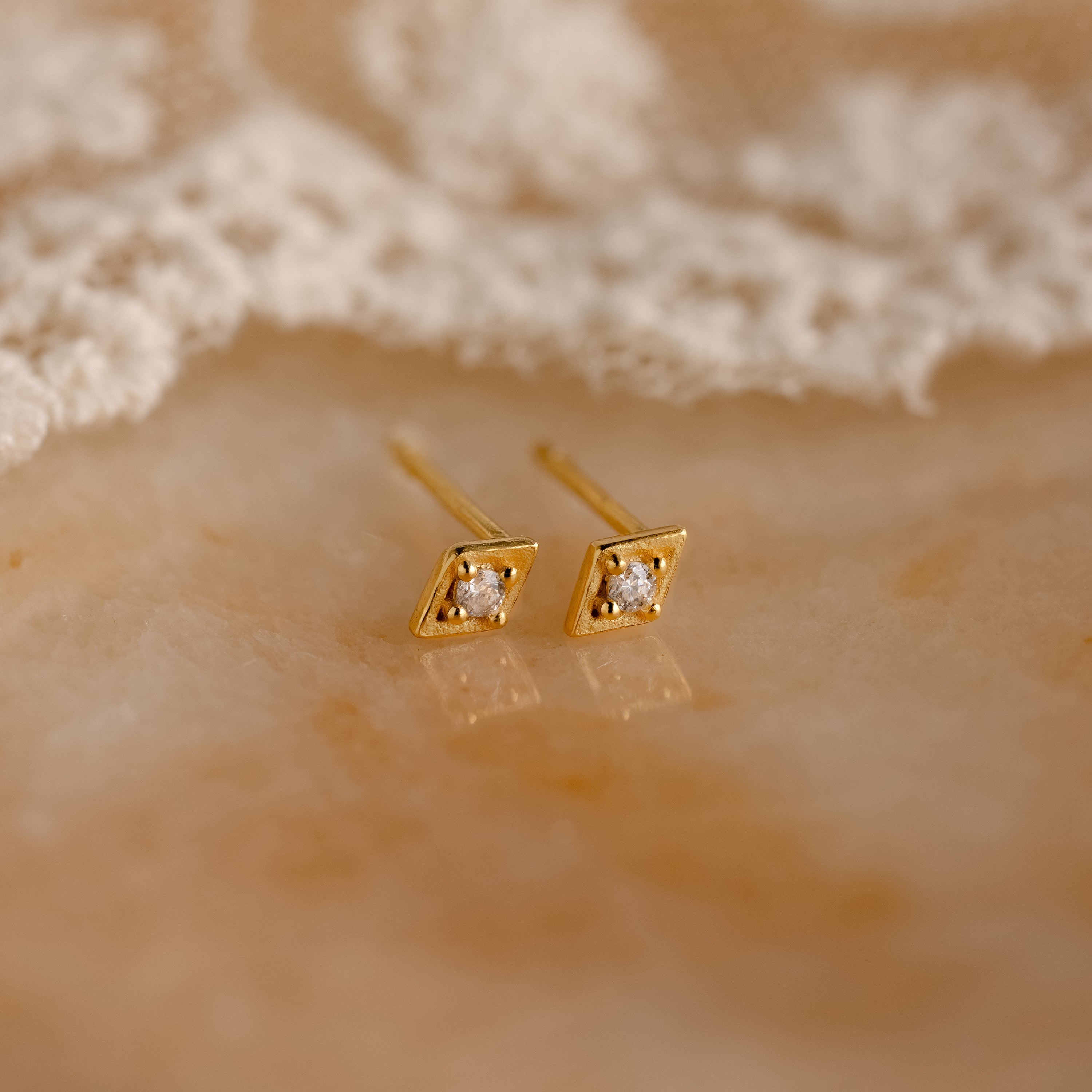 Ericka Diamond Studs are vintage-inspired, gold geometric earrings with small gemstones. These minimalist diamond studs are displayed elegantly on a marble surface next to delicate lace fabric.