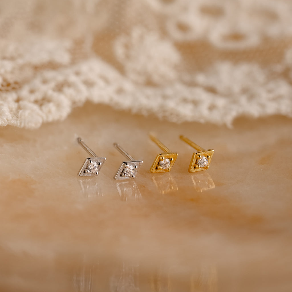 Two pairs of Ericka Diamond Studs, one in silver and one in gold, are displayed on a marble surface with lace in the background.