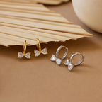 Diamond Bow Huggies in Silver, sparkling hoop bowtie earrings with clear gemstones, displayed on a beige background with a palm leaf.