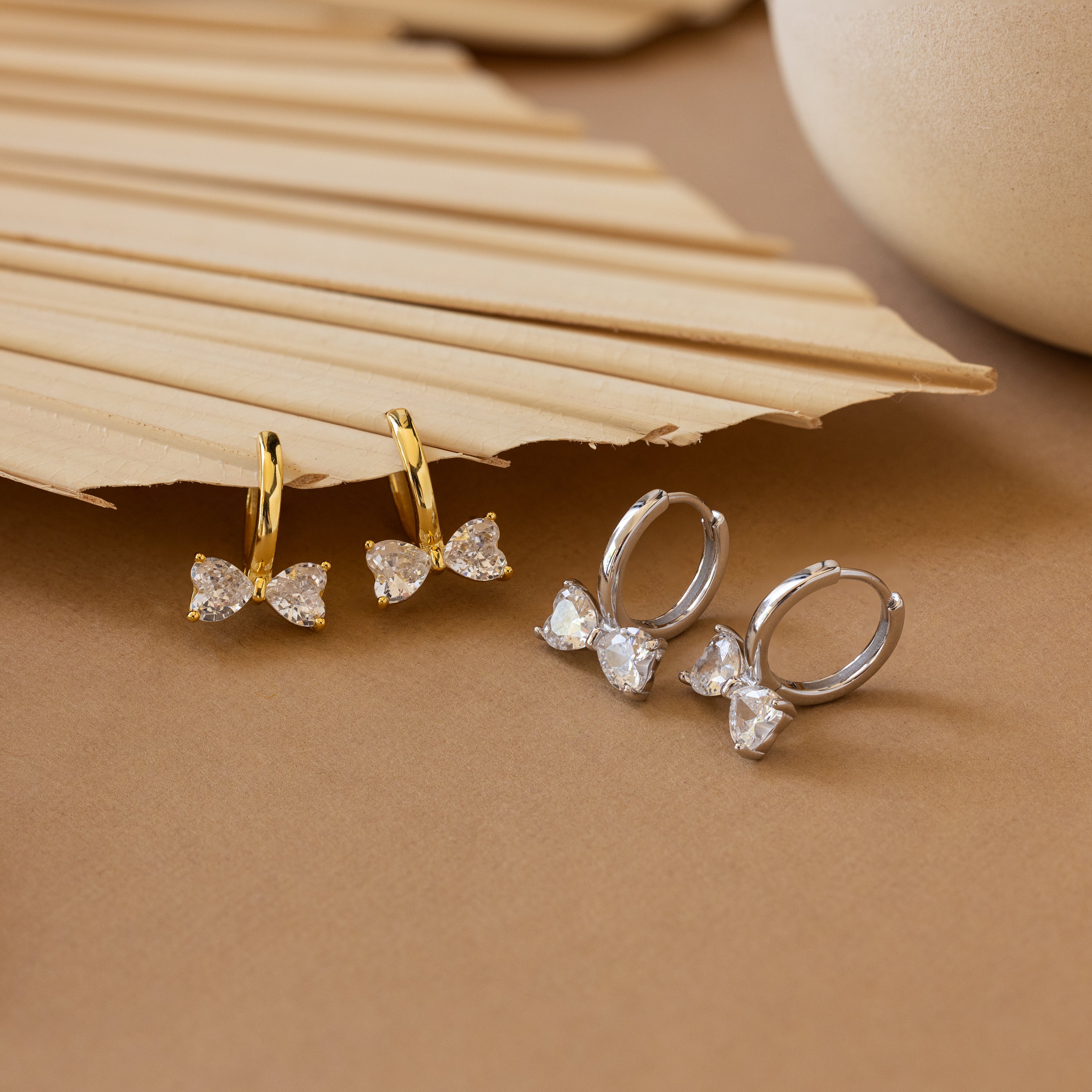 Diamond Bow Huggies: Bowtie earrings with clear stones, inspired by the iconic design, shown on a beige surface with a palm leaf accent.