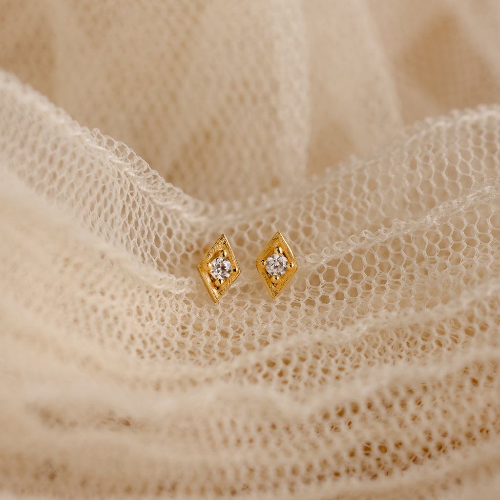 The Ericka Diamond Studs, featuring a gold geometric design with small central gems, rest on soft cream lace, capturing vintage-inspired minimalist elegance.