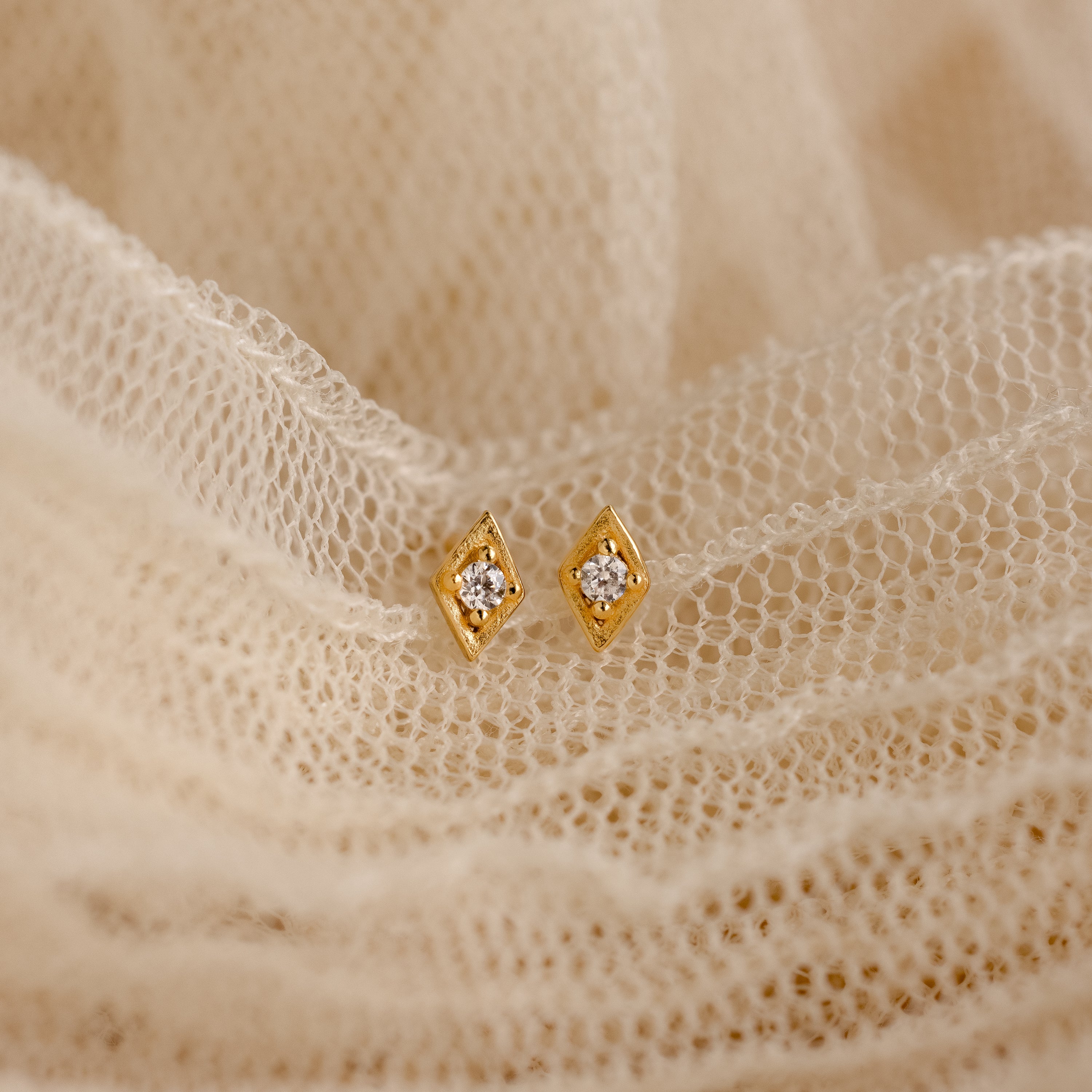 The Ericka Diamond Studs, featuring a gold geometric design with small central gems, rest on soft cream lace, capturing vintage-inspired minimalist elegance.