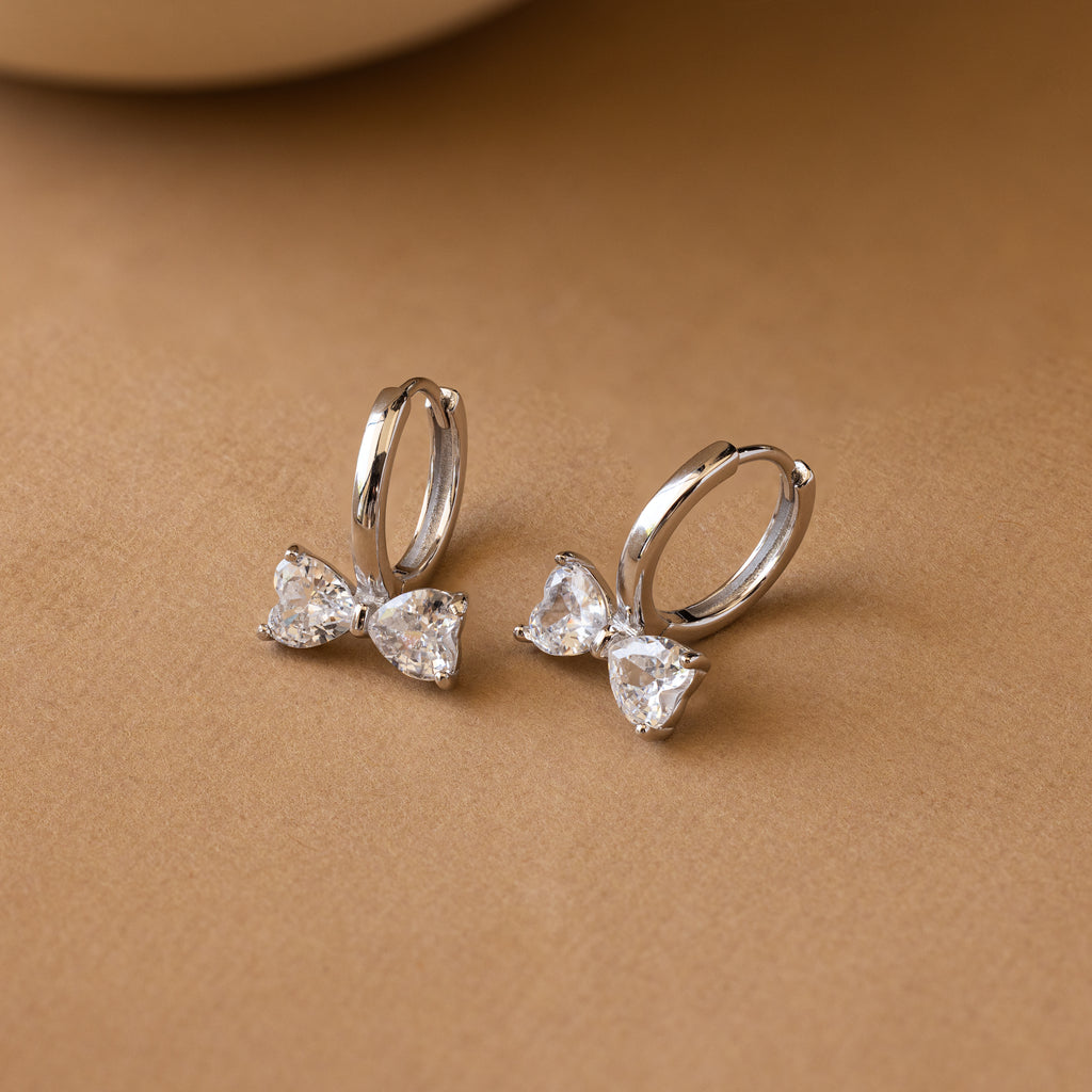 Diamond Bow Huggies: silver hoop earrings featuring bow-shaped clear gemstones. Perfect for adding chic elegance to any look for lovers of stylish bowtie earrings.