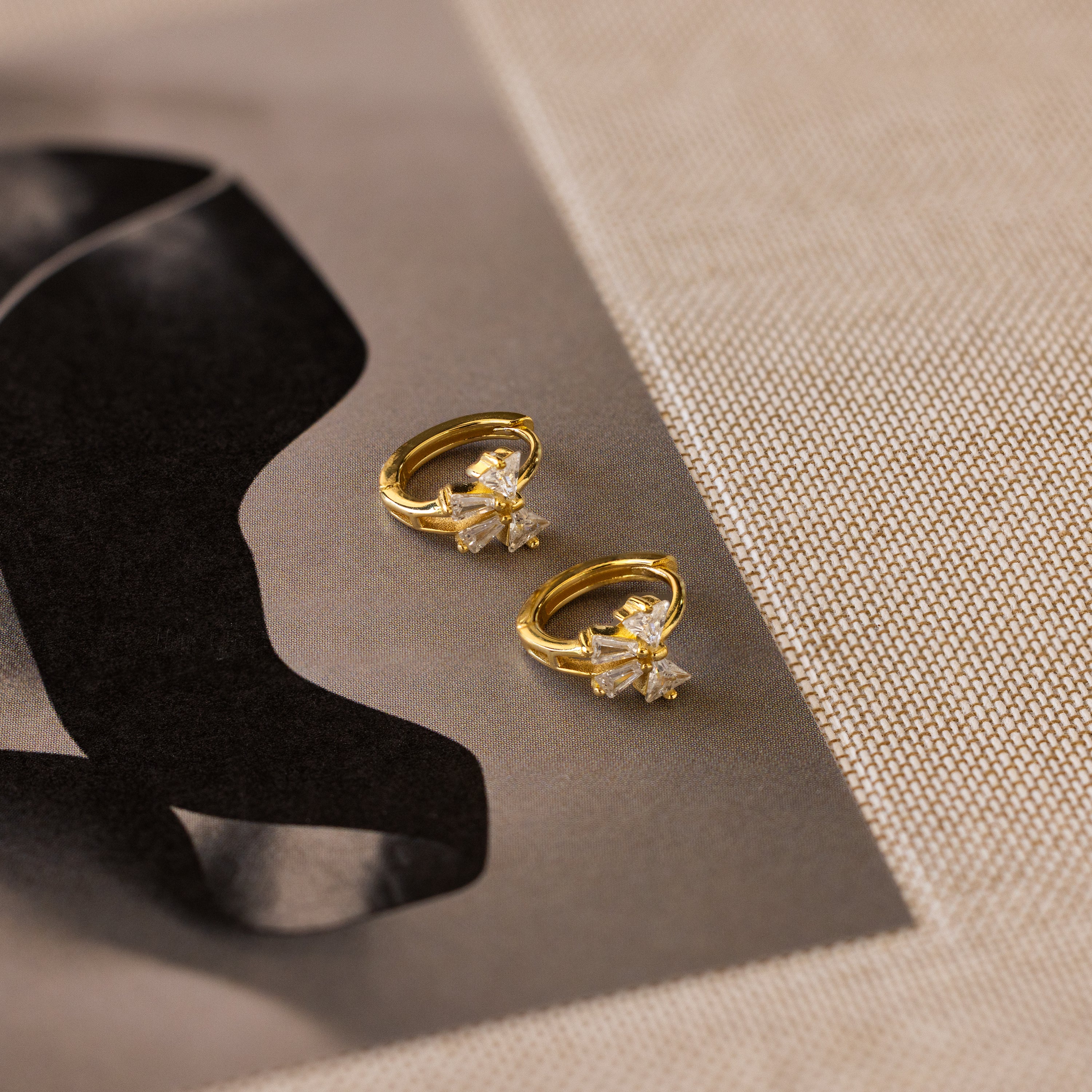 Diamond Ribbon Bow Huggies in 18K Gold are displayed on a textured surface next to abstract black artwork.