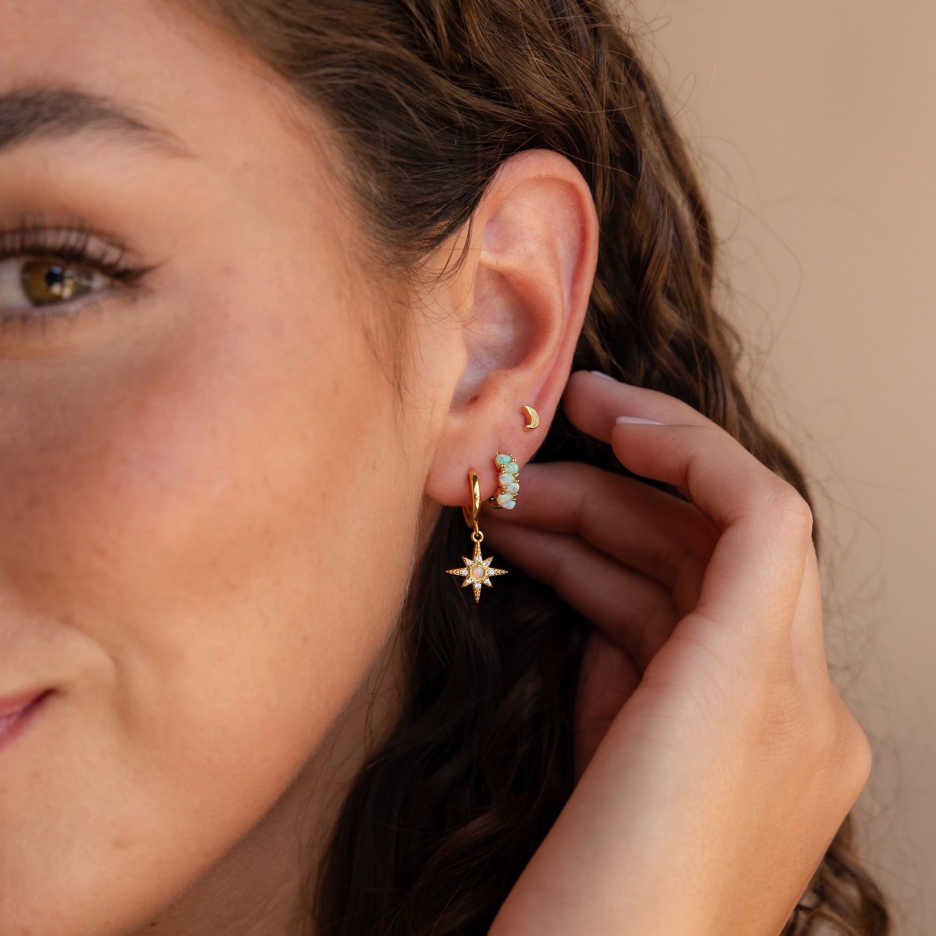 Close-up of a woman’s ear wearing Ombre Opal Huggies, a star dangle earring, and a small crescent moon earring.