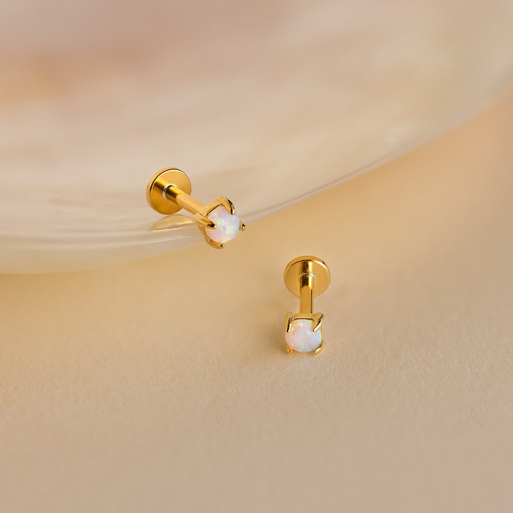 Two Opal Flat Back Studs in 18K Gold with small round opal stones rest on a soft beige surface near a shell—perfect dainty earrings for everyday elegance.