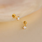 Two Opal Flat Back Studs in 18K Gold with small round opal stones rest on a soft beige surface near a shell—perfect dainty earrings for everyday elegance.