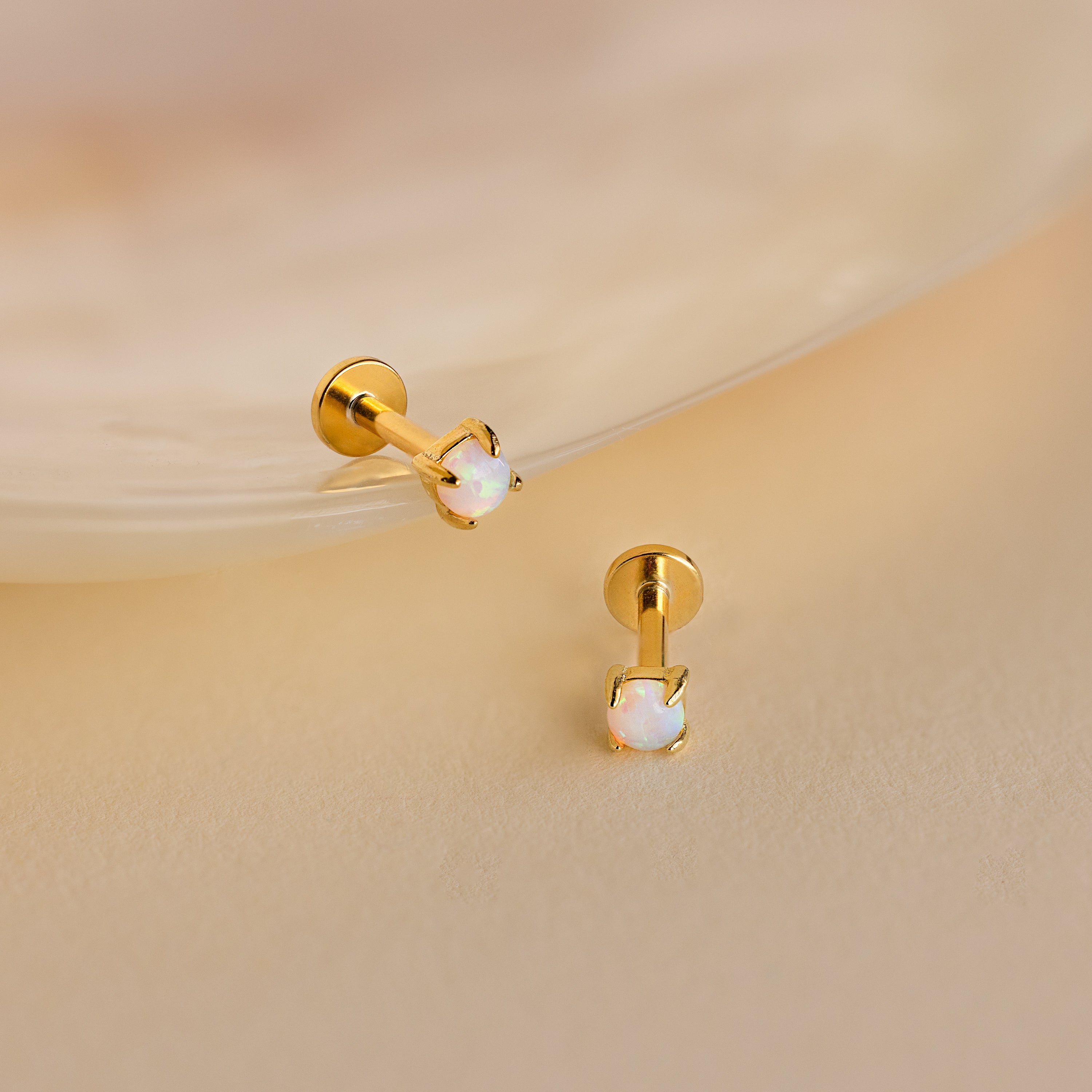 Two Opal Flat Back Studs in 18K Gold with small round opal stones rest on a soft beige surface near a shell—perfect dainty earrings for everyday elegance.