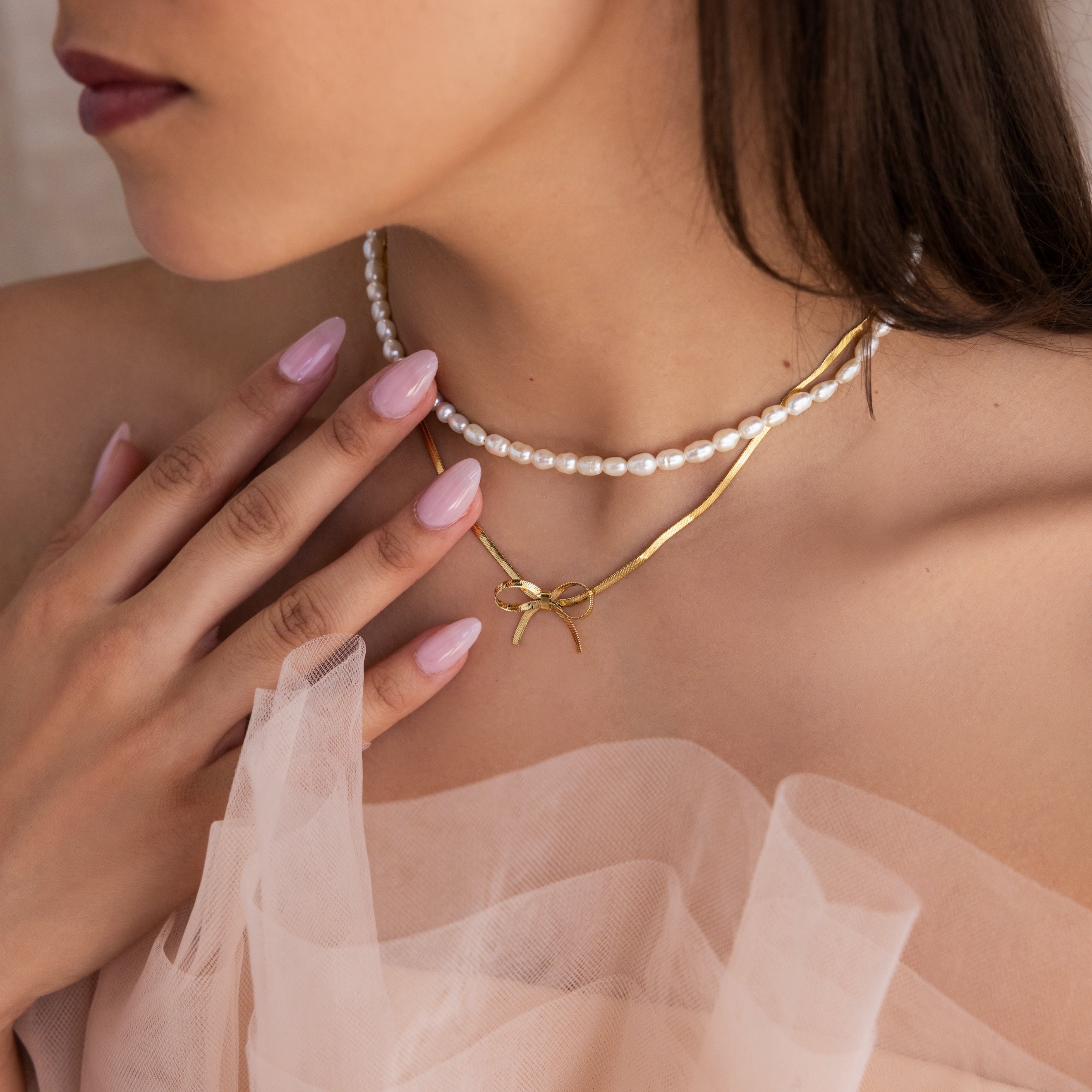 A woman with manicured nails and a sheer tulle top wears the Herringbone Bow Necklace in 18K Gold and a pearl necklace for a chic, coquette style.