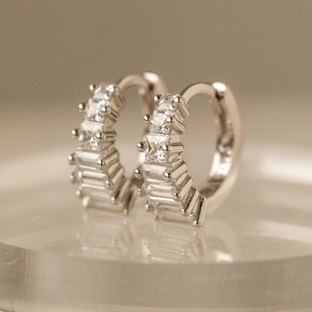 Close-up view of silver Art Deco Huggie earrings, intricately designed with a mix of brilliant-cut and baguette-cut cubic zirconia stones set in a geometric stair-step pattern. The earrings are elegantly perched on a clear, rounded display stand, highlighting their structured silhouette and radiant sparkle under soft lighting.