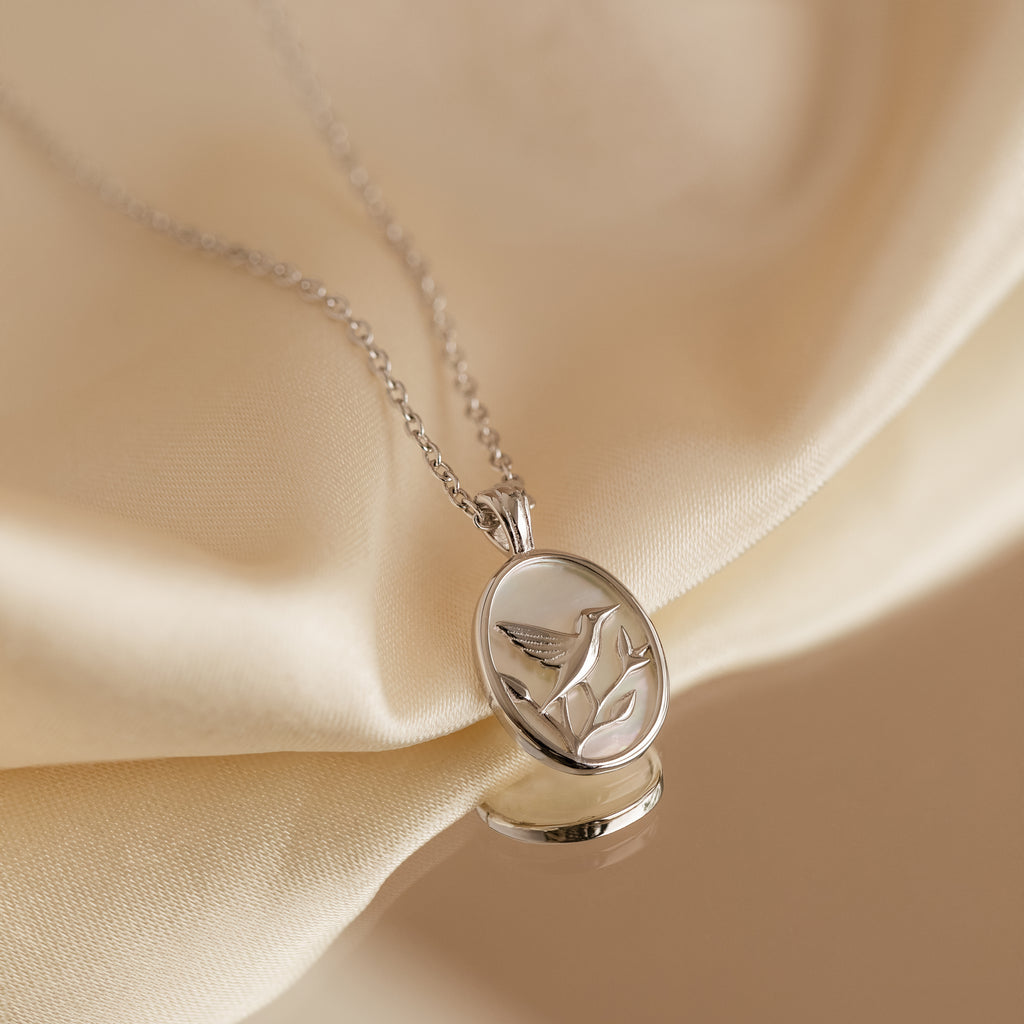 The Pearl Hummingbird Necklace in Sterling Silver features a delicate design displayed on soft cream fabric—an elegant addition to any minimalist jewelry collection.